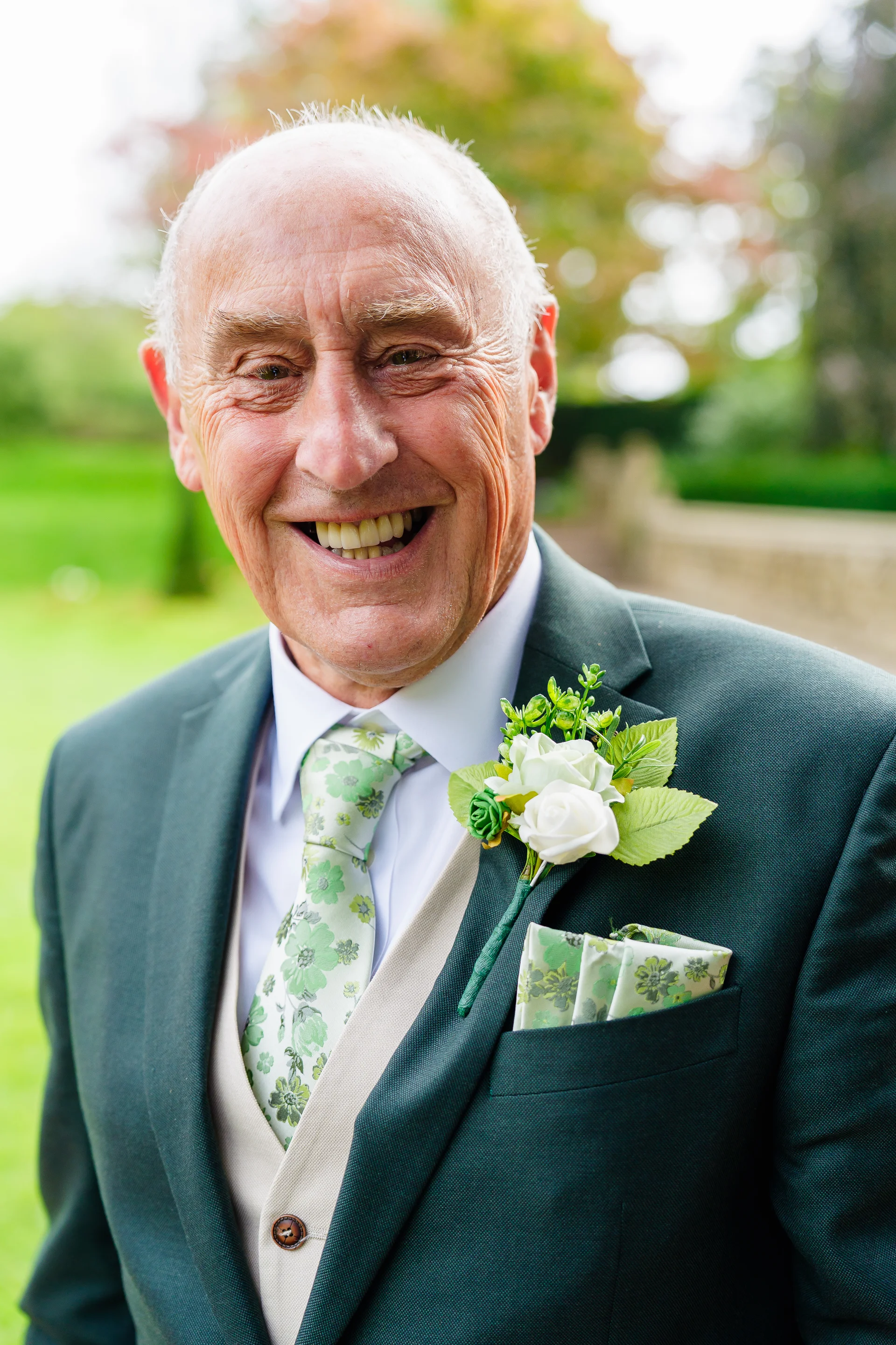 An elderly gentleman smiles warmly at the camera, wearing a dark green suit, a floral patterned tie, and a matching white rose boutonniere pinned to his lapel.