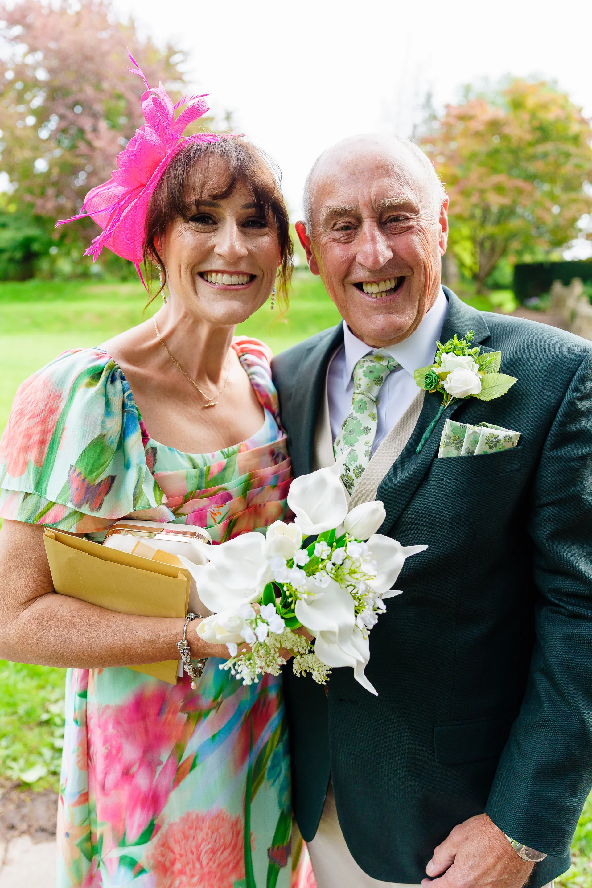 A smiling older couple attending an outdoor wedding, with the woman wearing a floral dress and a bright pink fascinator while holding an envelope, and the man wearing a green suit with a white floral buttonhole.