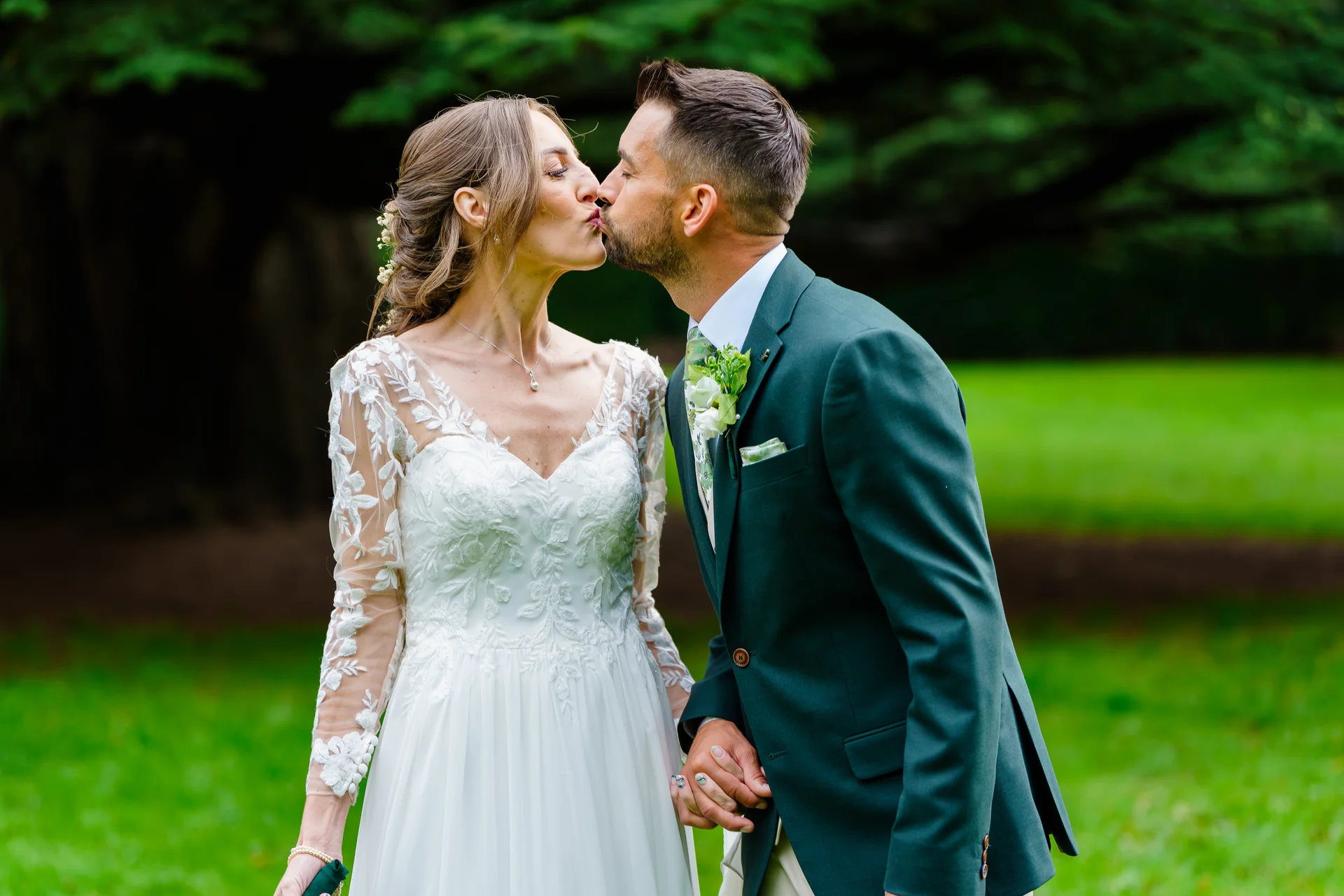 The newly married couple share a tender kiss outdoors against a lush green woodland backdrop, with the bride wearing a lace long-sleeve gown and the groom in a dark green suit.