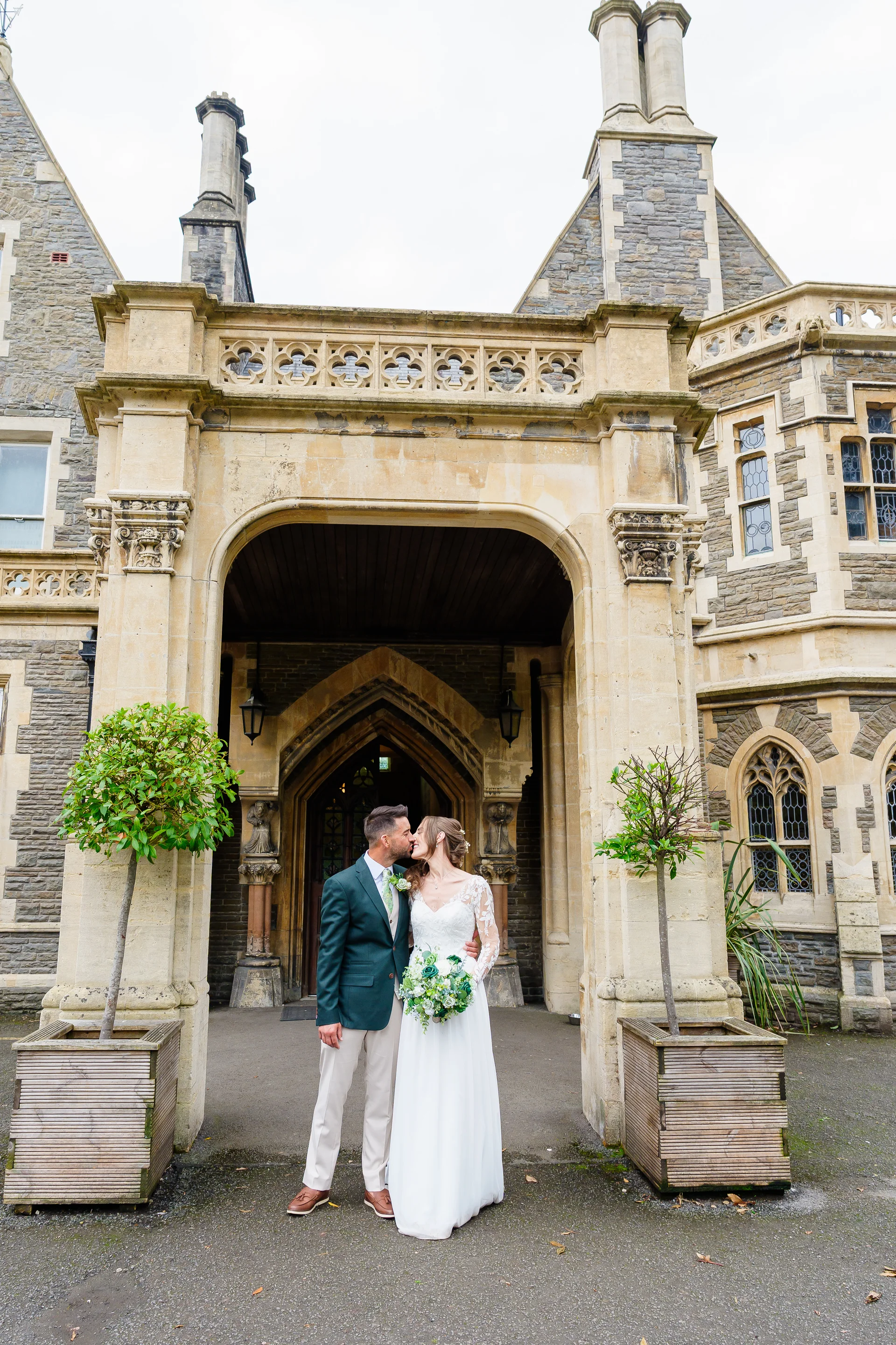 A newlywed couple shares a tender kiss beneath the grand stone entrance arch of a historic country house wedding venue.