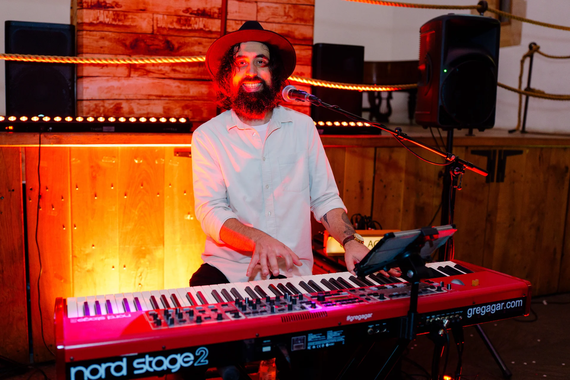 A bearded male musician wearing a hat and light shirt plays a red keyboard on stage during a rustic barn wedding reception.