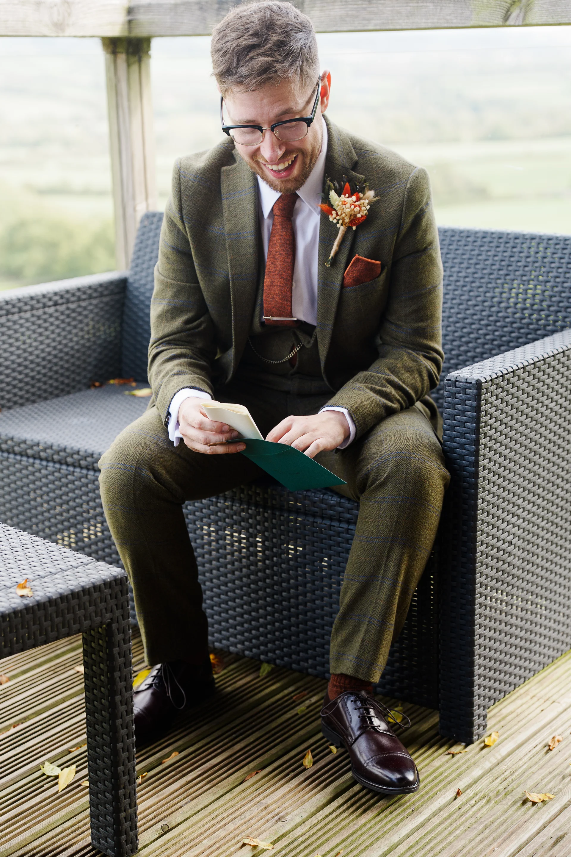 A smiling groom wearing a green tweed suit and rust-coloured tie sits on an outdoor rattan sofa while reading a heartfelt letter.