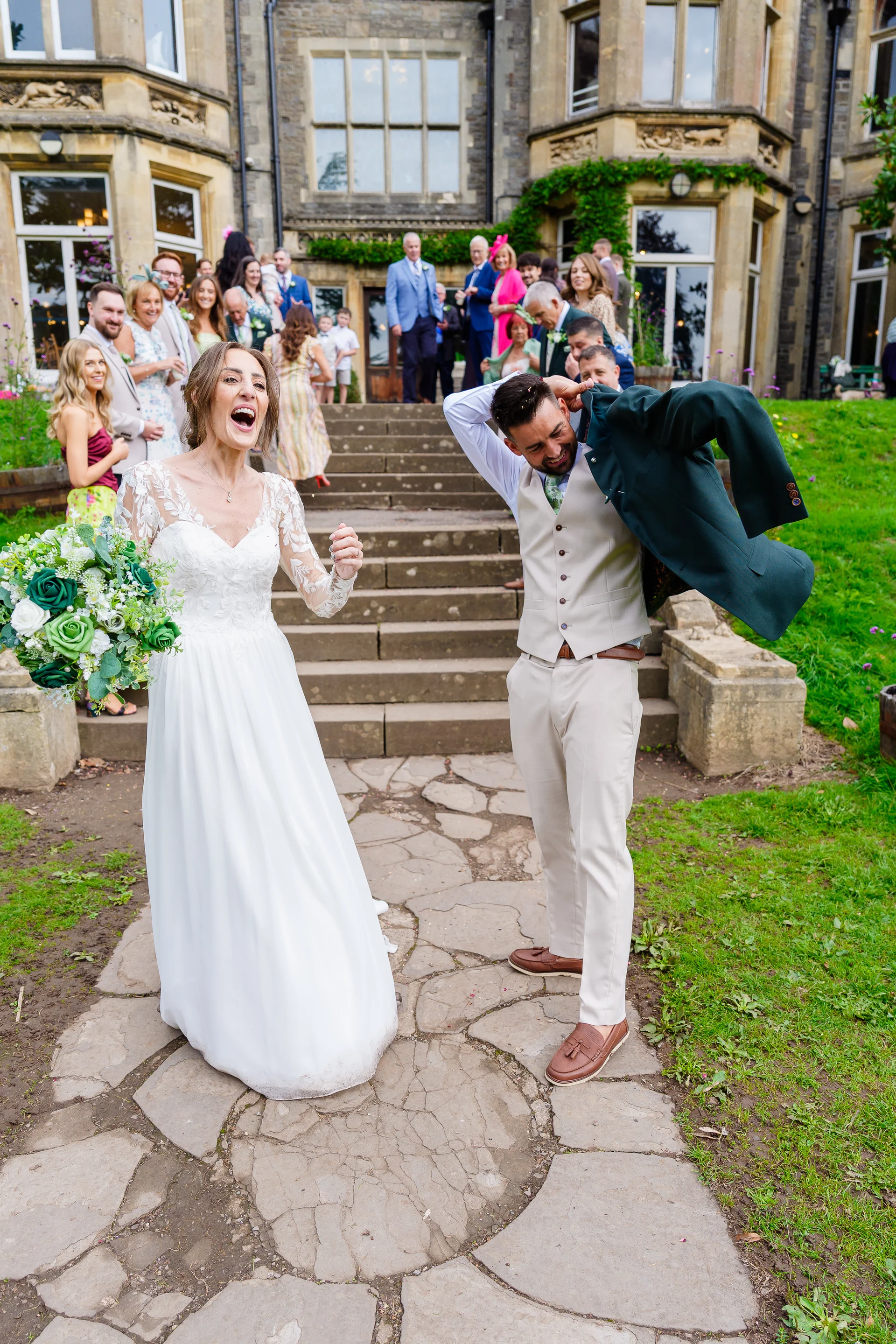 A joyous bride holding a green and white bouquet and a groom slipping into his jacket laugh ecstatically on stone steps outside a grand country house as guests cheer in the background.