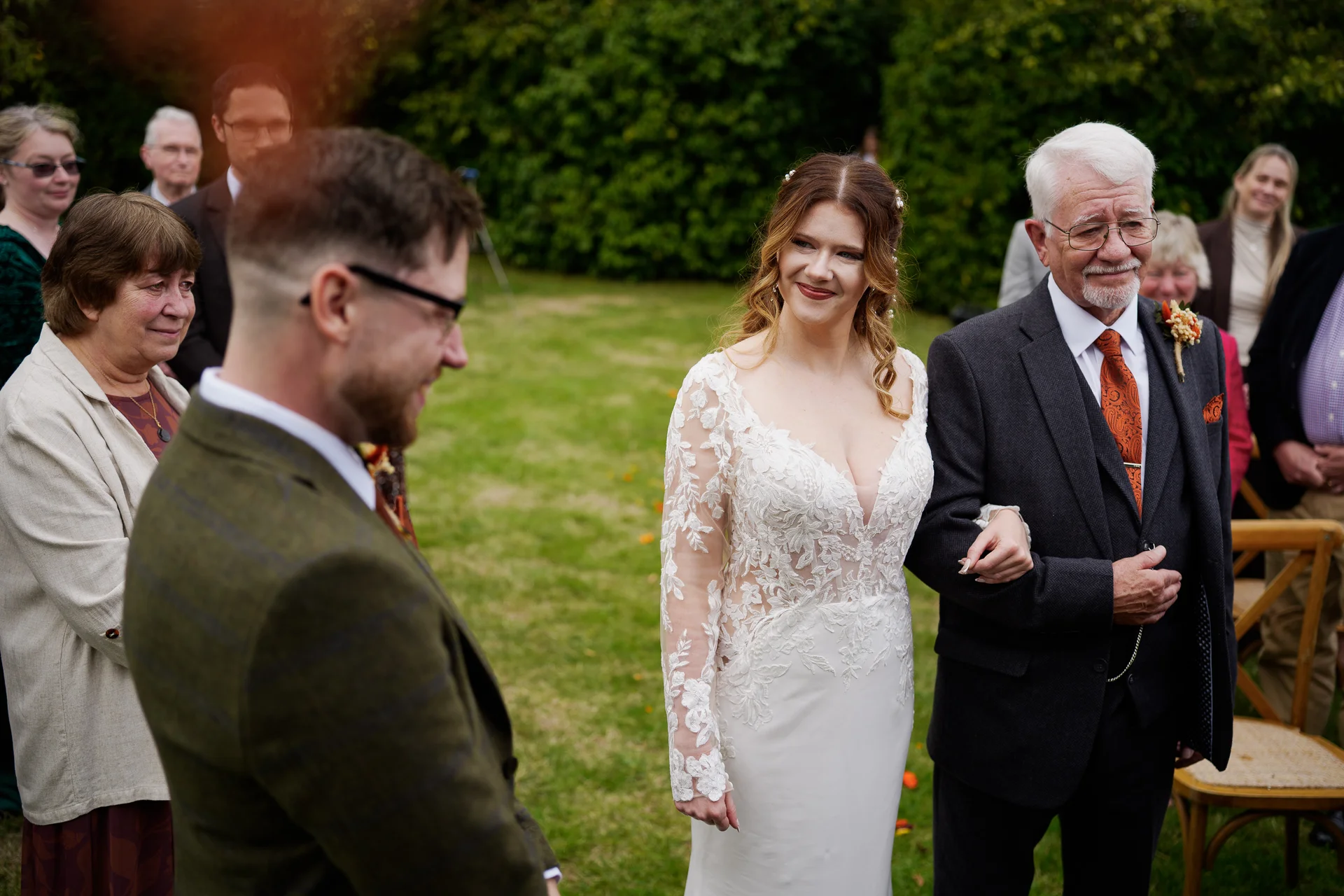 A smiling bride walks down the aisle arm in arm with her father, looking lovingly towards the waiting groom during an outdoor ceremony.