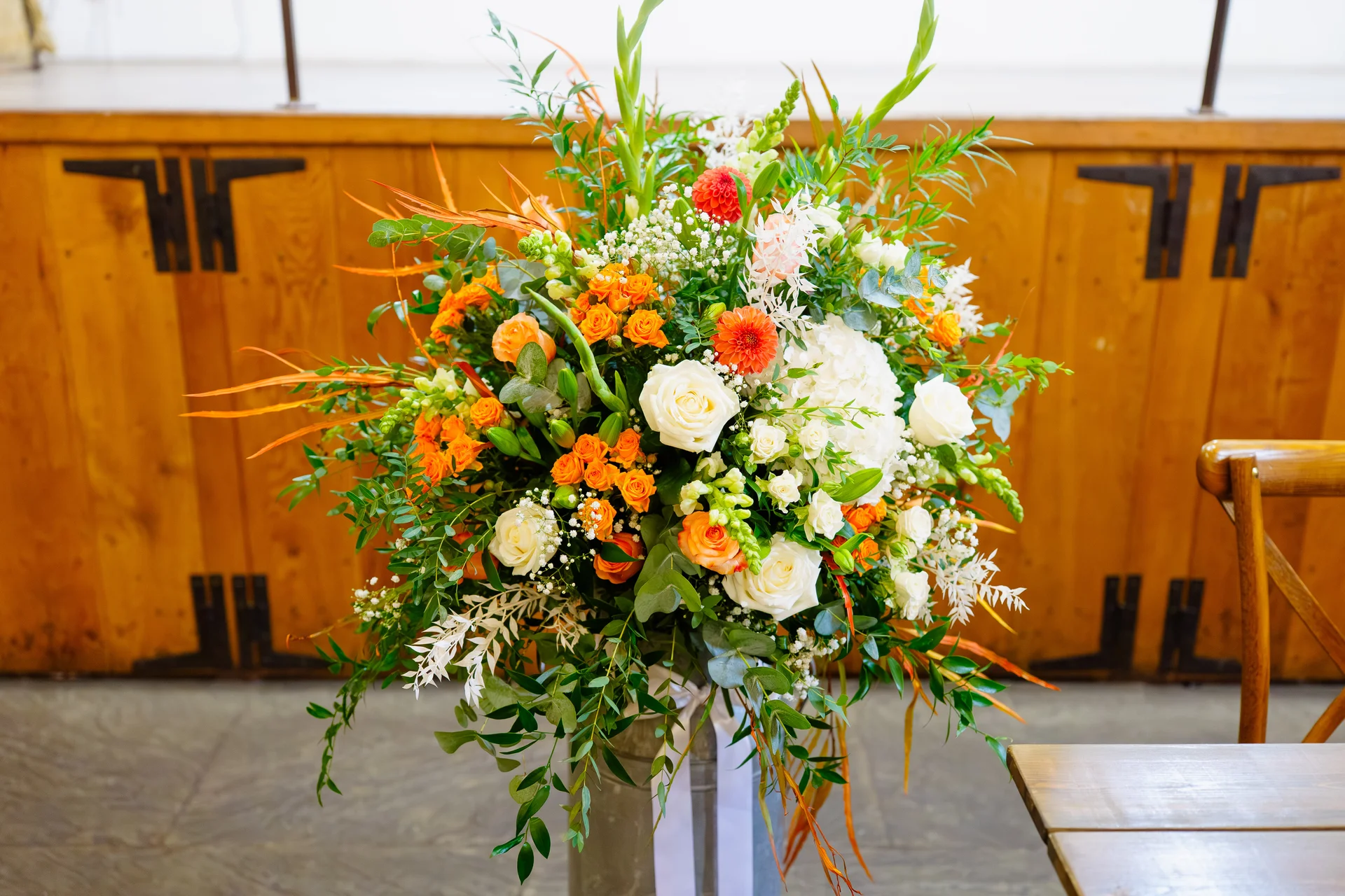 A large, vibrant floral pedestal arrangement featuring white roses, hydrangeas, and bright orange accents stands in front of a rustic wooden panelled wall.