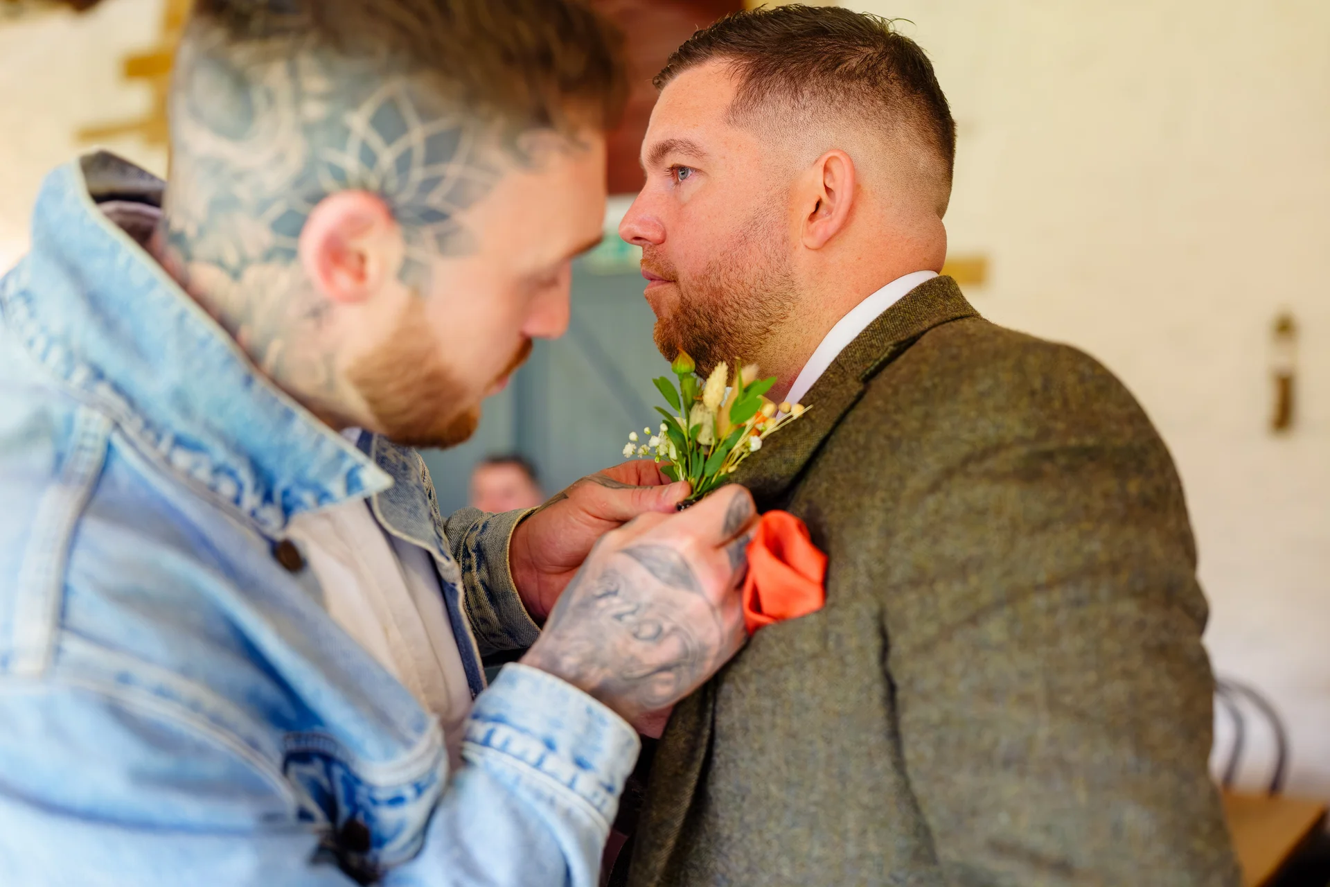 A heavily tattooed person carefully pins a floral buttonhole onto the lapel of a groom wearing a tweed suit.