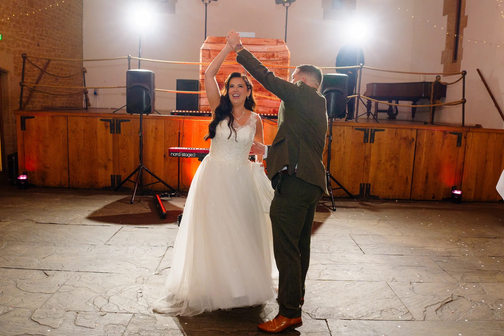 A newlywed couple joyfully shares their first dance on a stone floor inside a rustic stone barn with wooden paneling and stage equipment in the background.