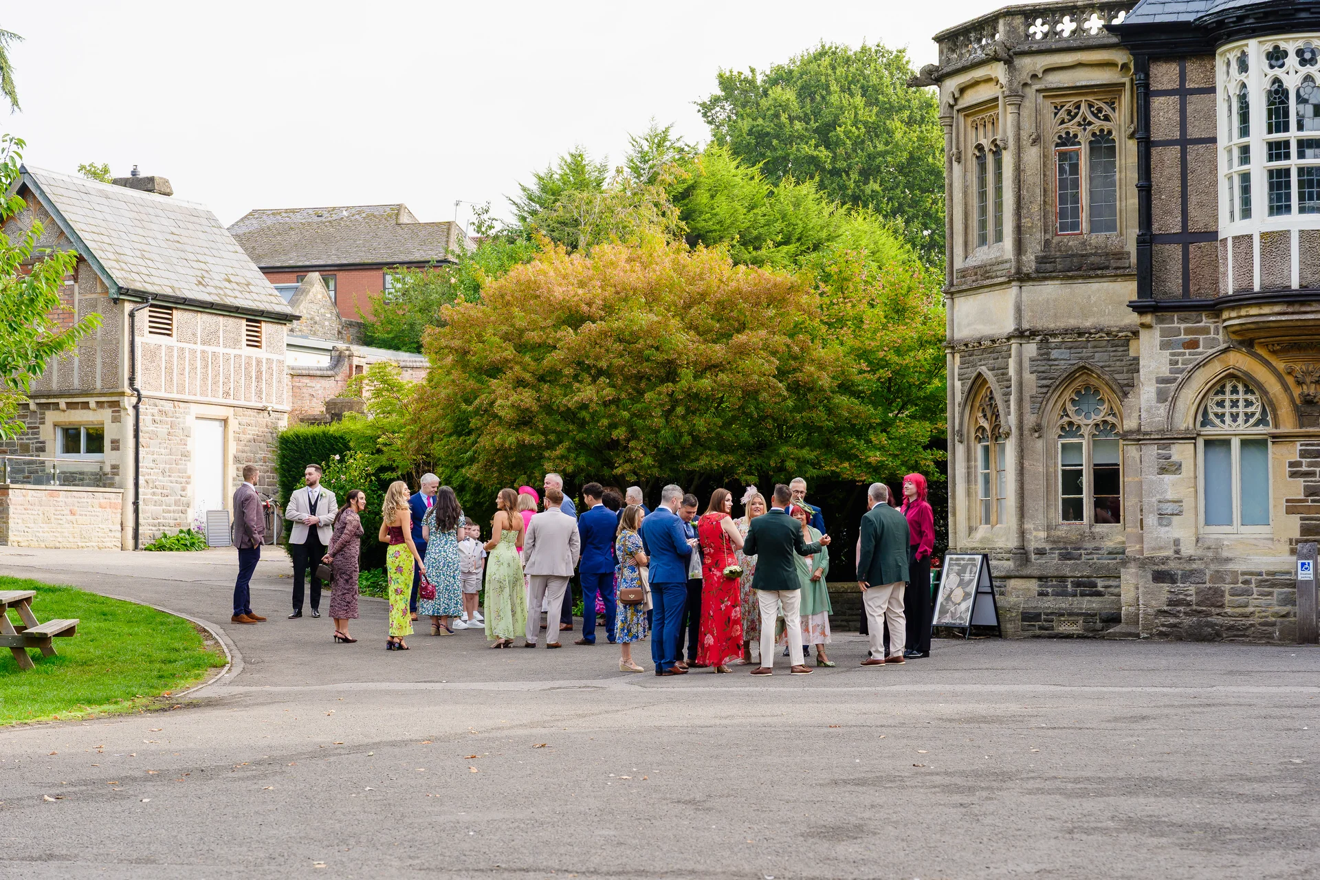 Wedding guests mingle and chat outside the stone walls and gothic windows of a historic country house venue during the afternoon reception.