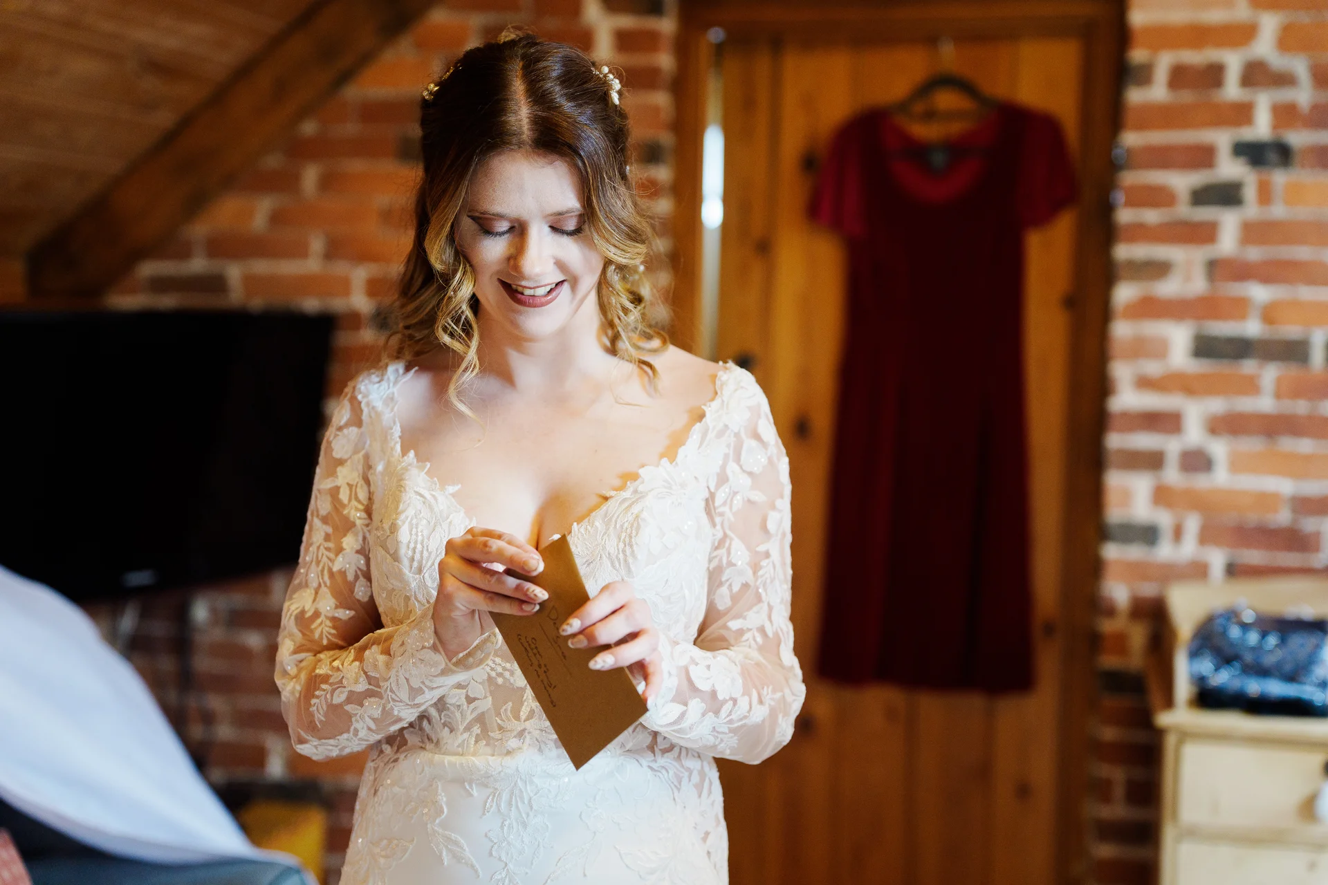 A smiling bride in a lace long-sleeve gown reads a heartfelt letter during bridal preparations inside a rustic room with exposed brick walls.