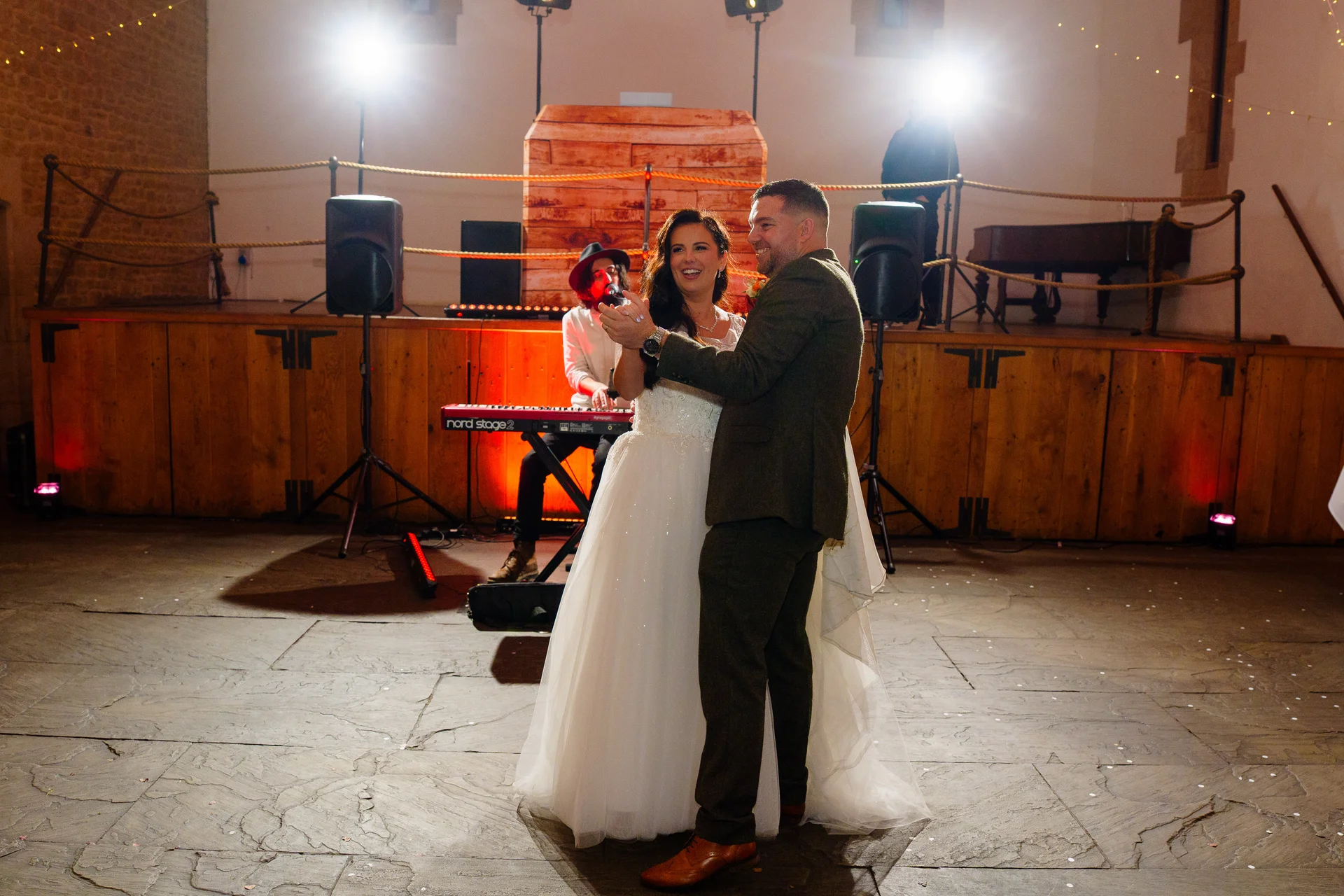 A newlywed couple shares their first dance on a stone flagstone floor in a rustic barn venue while a live band performs on a wooden stage in the background.
