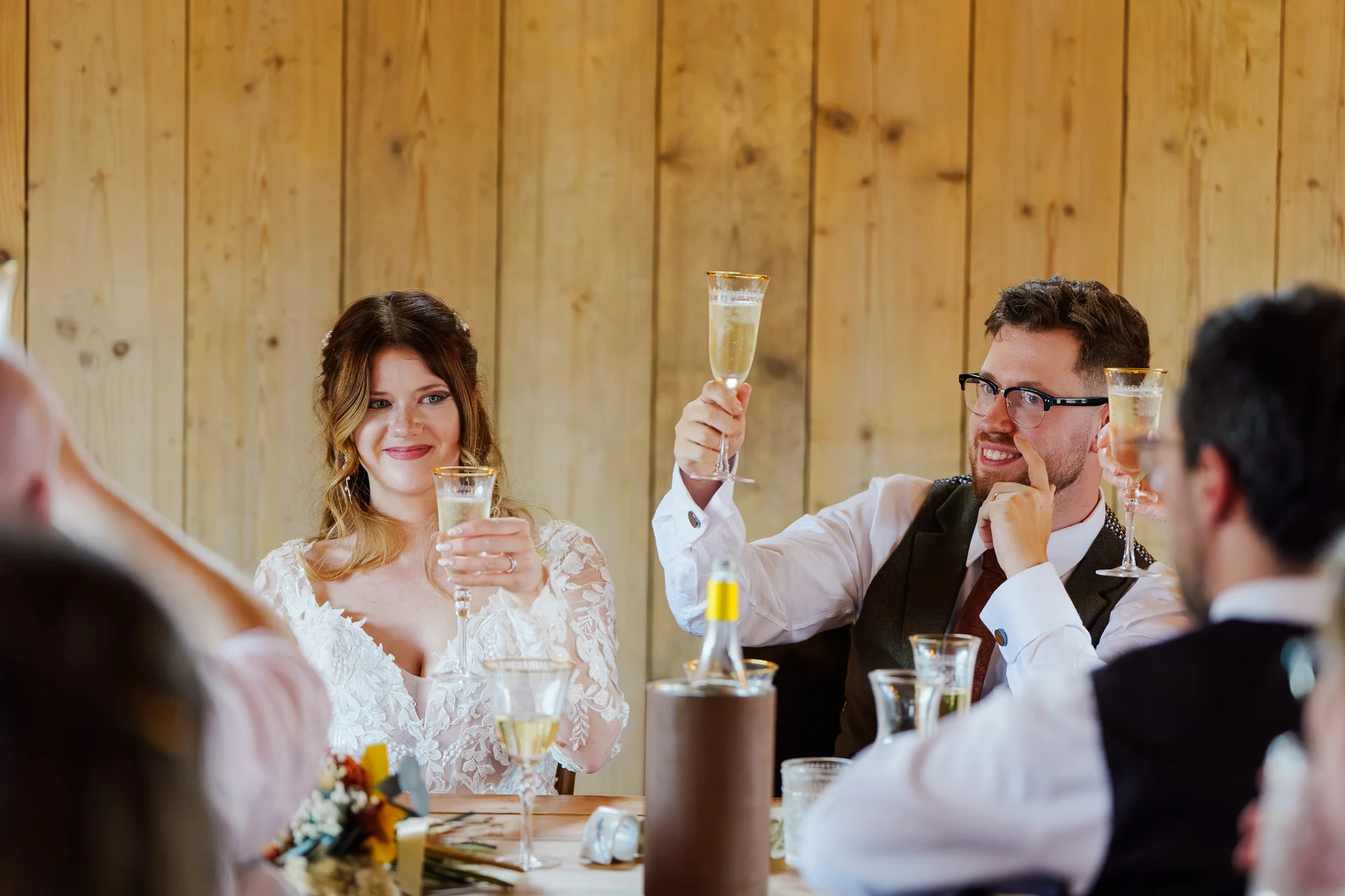 A smiling bride and groom raise their champagne flutes for a toast during the wedding speeches inside a rustic barn venue.