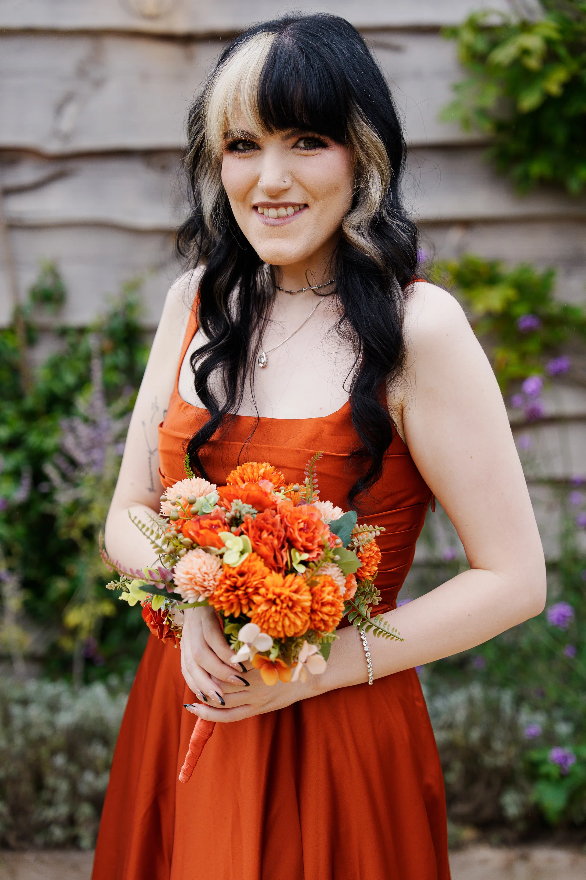 A smiling bride wearing a burnt orange dress and holding a vibrant autumn-toned bouquet stands outdoors against a rustic wooden background.