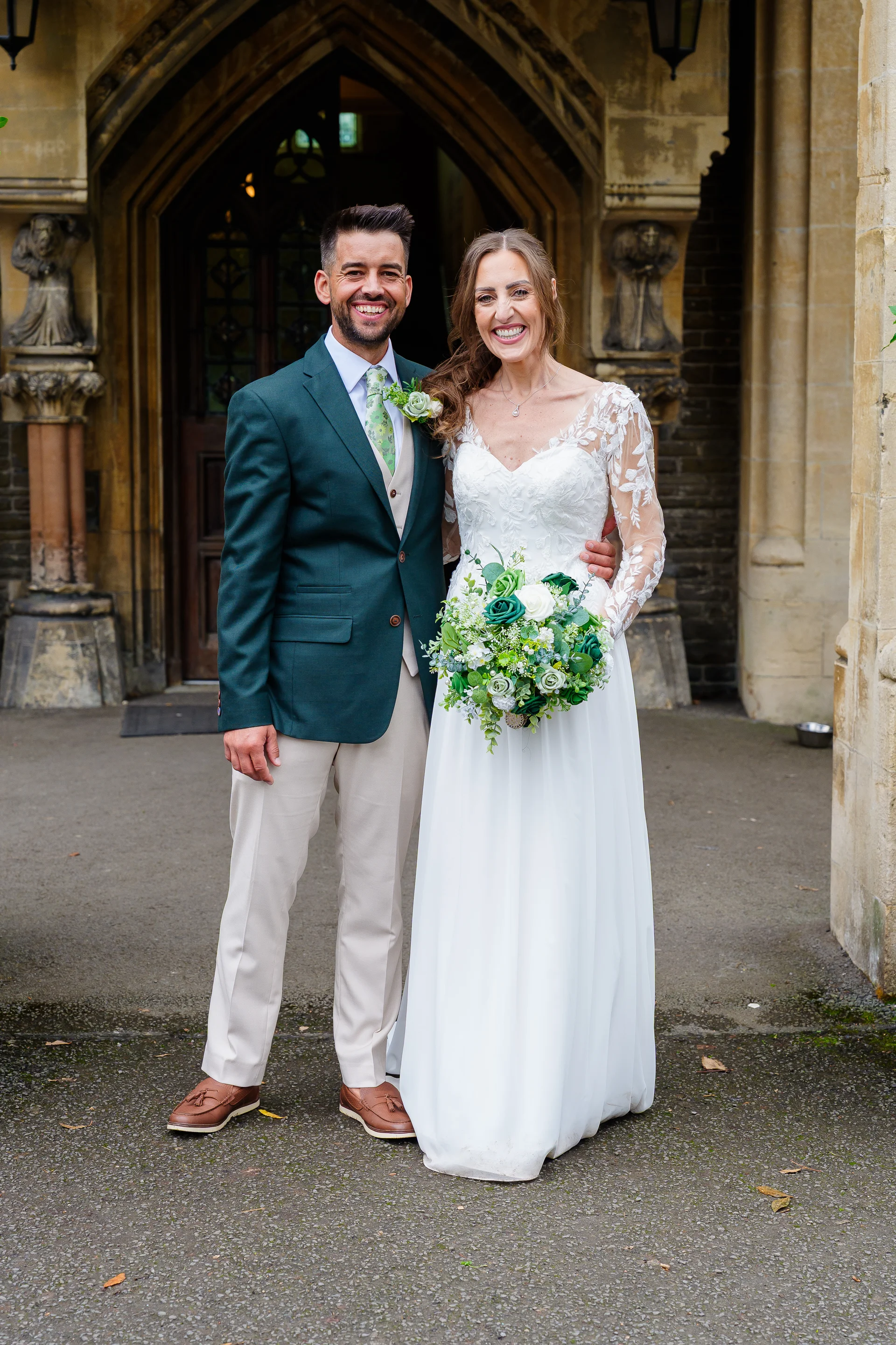 A smiling bride holding a green and white bouquet and a groom in a green suit pose happily outside an ornate stone church entrance.