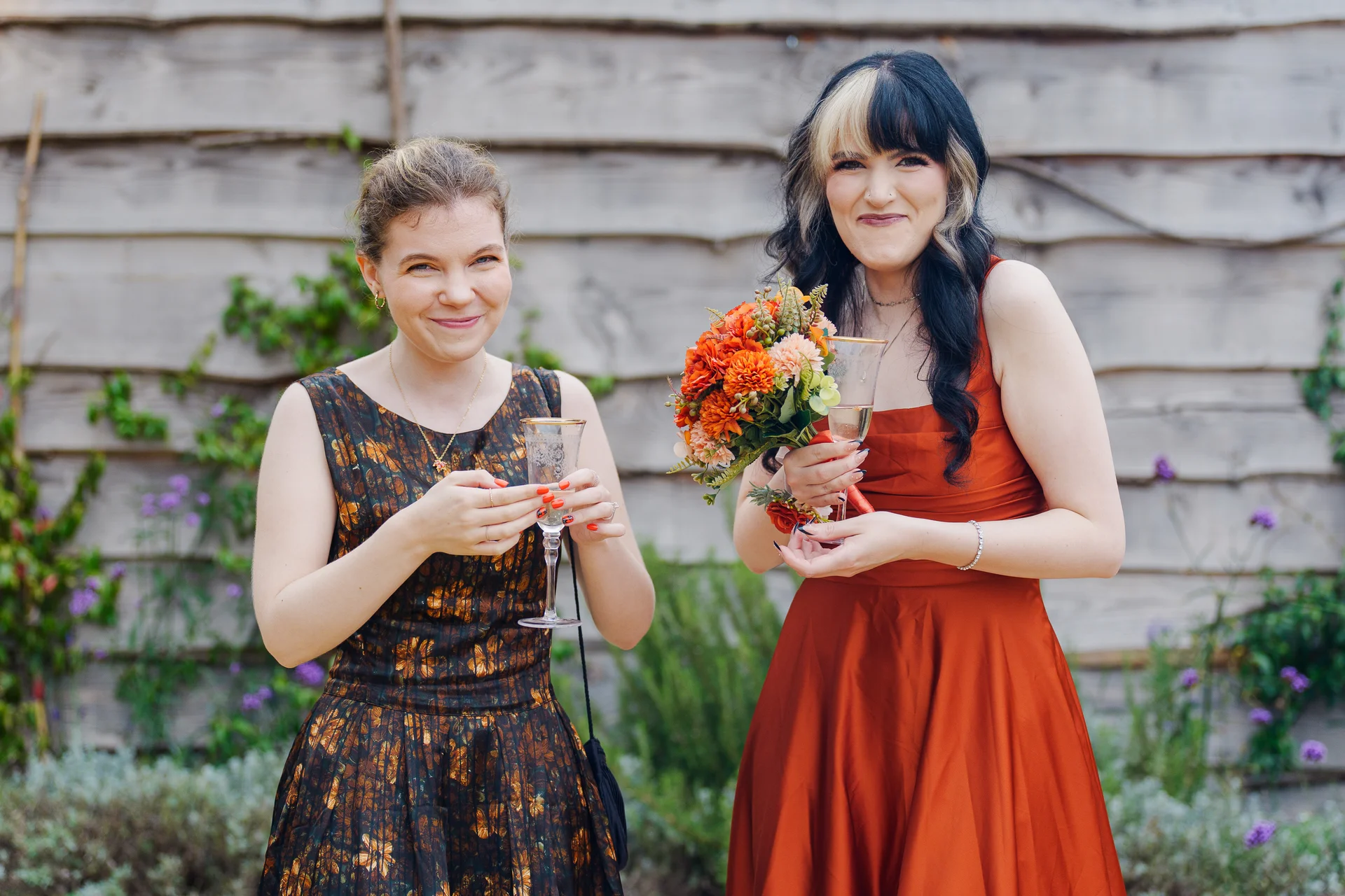Two smiling brides pose outdoors in front of a rustic wooden wall, holding champagne glasses and a vibrant orange and autumn-toned bridal bouquet.