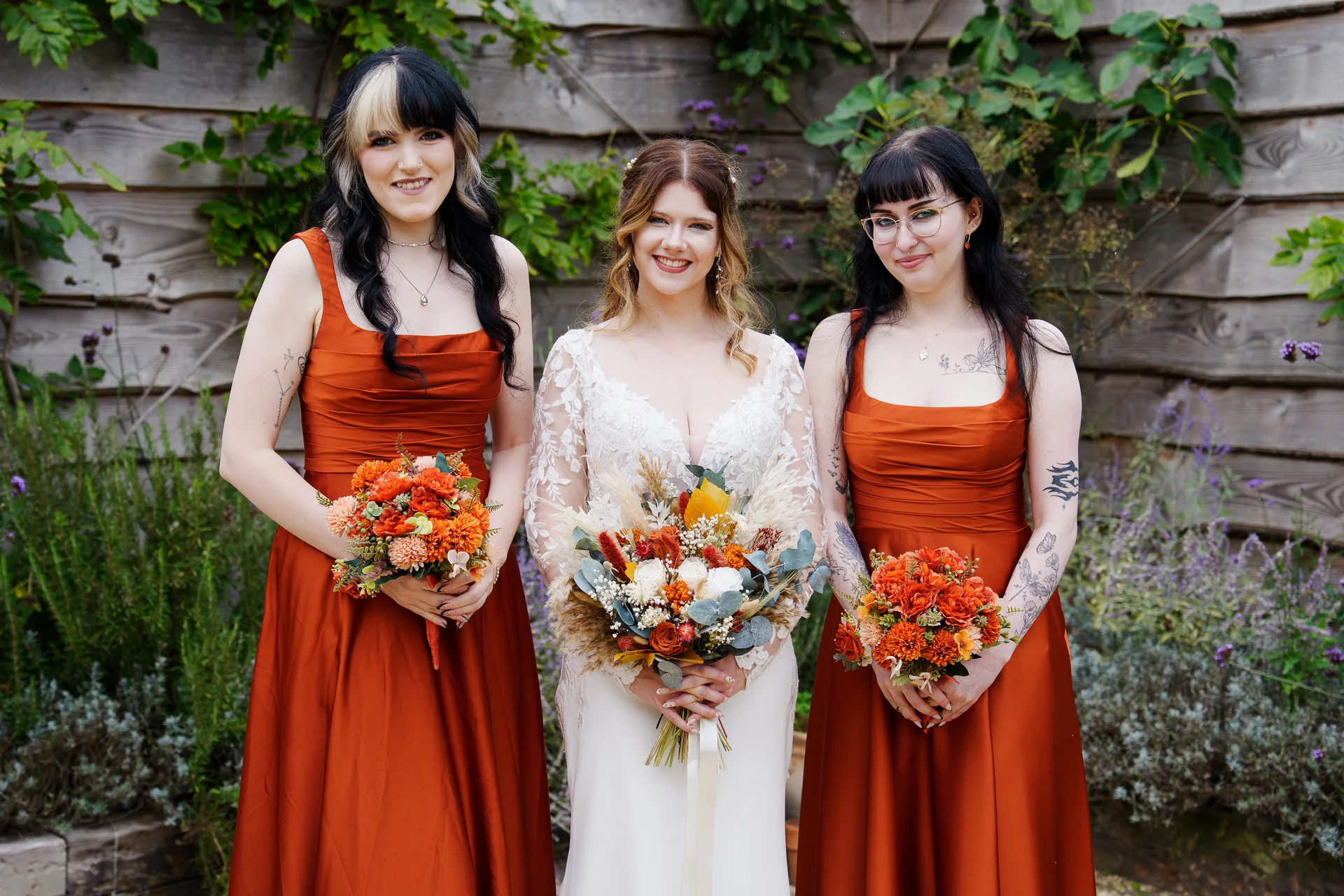 A smiling bride in a long-sleeved lace gown stands flanked by two bridesmaids in vibrant terracotta satin dresses, all holding autumnal orange bouquets in front of a rustic wooden fence.