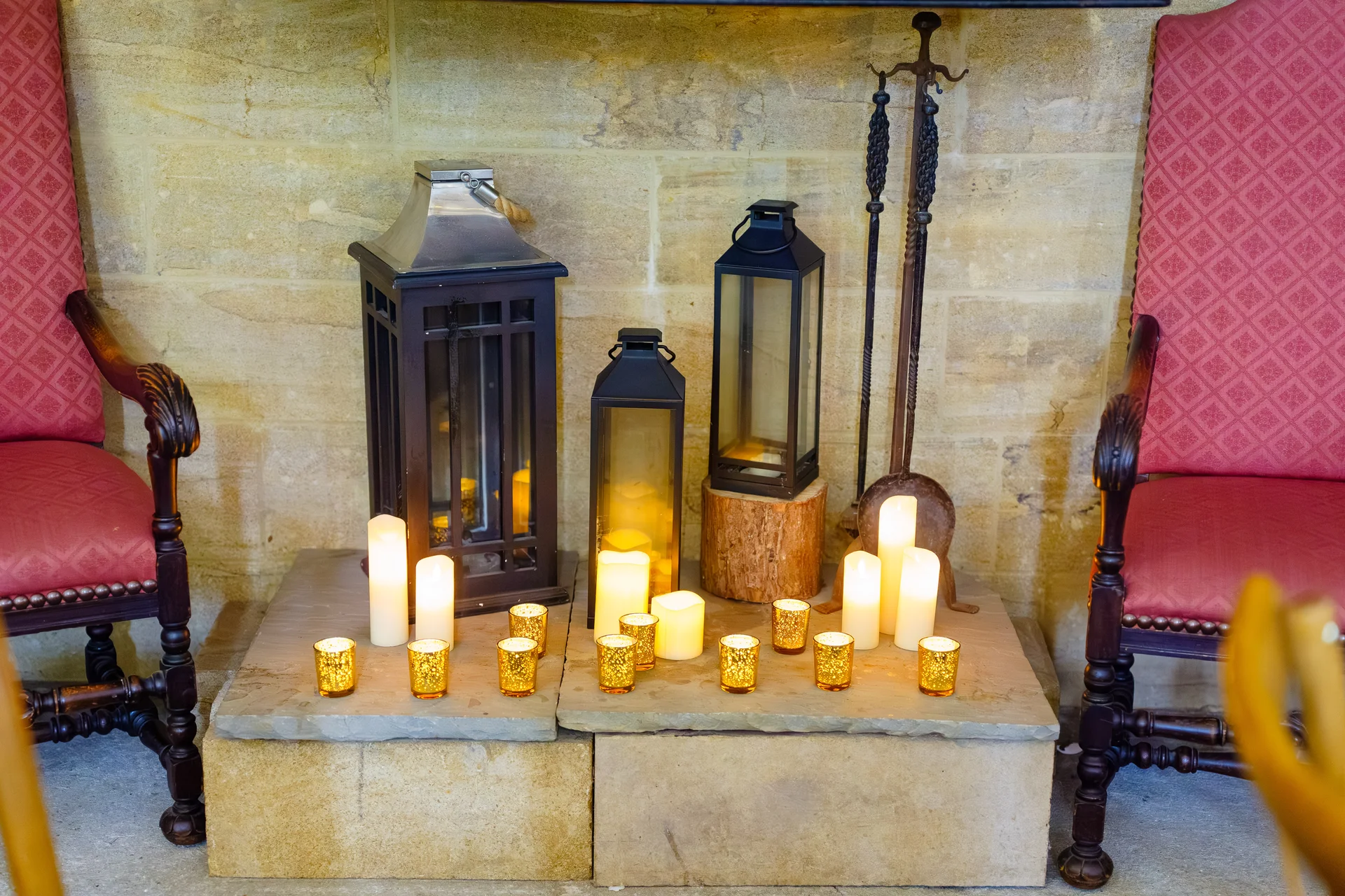 A glowing display of various black lanterns and gold votive candles arranged on a stone hearth between two red-upholstered armchairs.