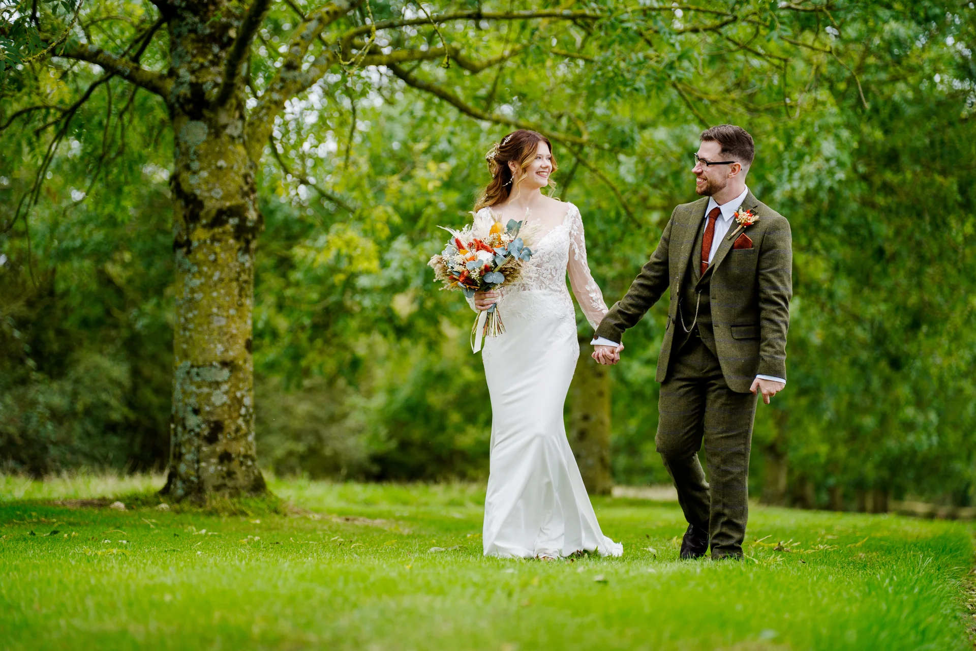 A newlywed couple walk hand in hand through a lush green woodland, laughing and looking at each other as the bride carries a colourful seasonal bouquet.