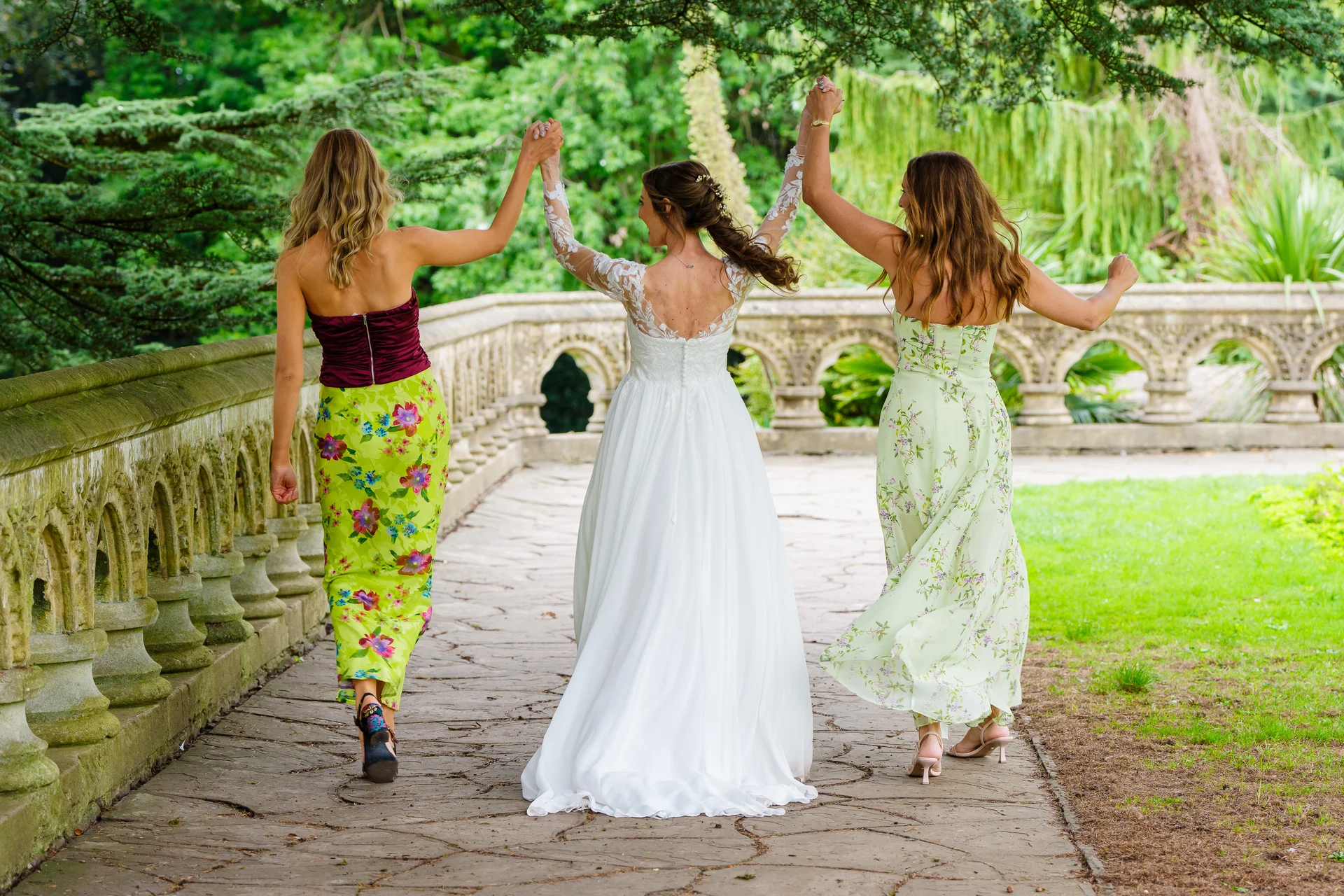 A bride in a lace-sleeved wedding gown and her two bridesmaids walk hand-in-hand along a stone bridge in a lush garden, viewed from behind.