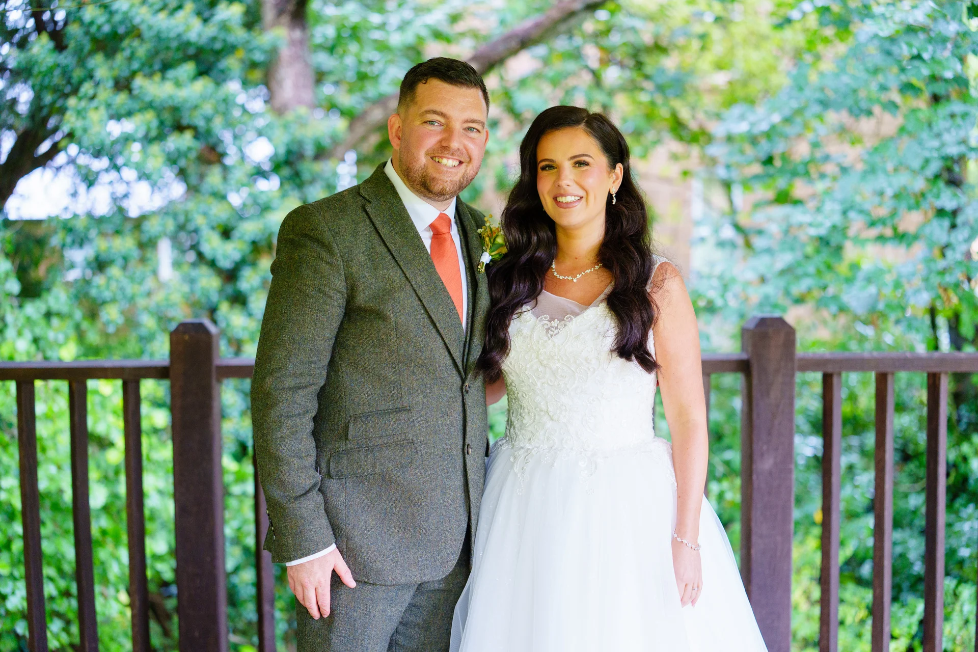 A smiling bride and groom pose together outdoors against a lush green backdrop, with the groom wearing a tweed suit and the bride in a lace gown.