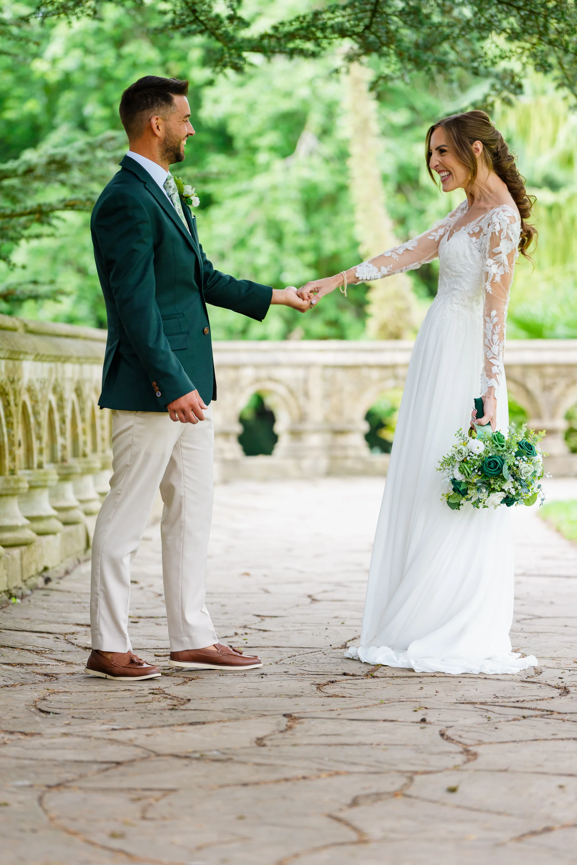 A newlywed couple holds hands and laughs together while walking across a stone bridge outdoors on their wedding day.