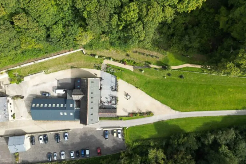 An overhead drone shot captures a wedding venue nestled beside a lush green woodland with a paved courtyard and parked cars.