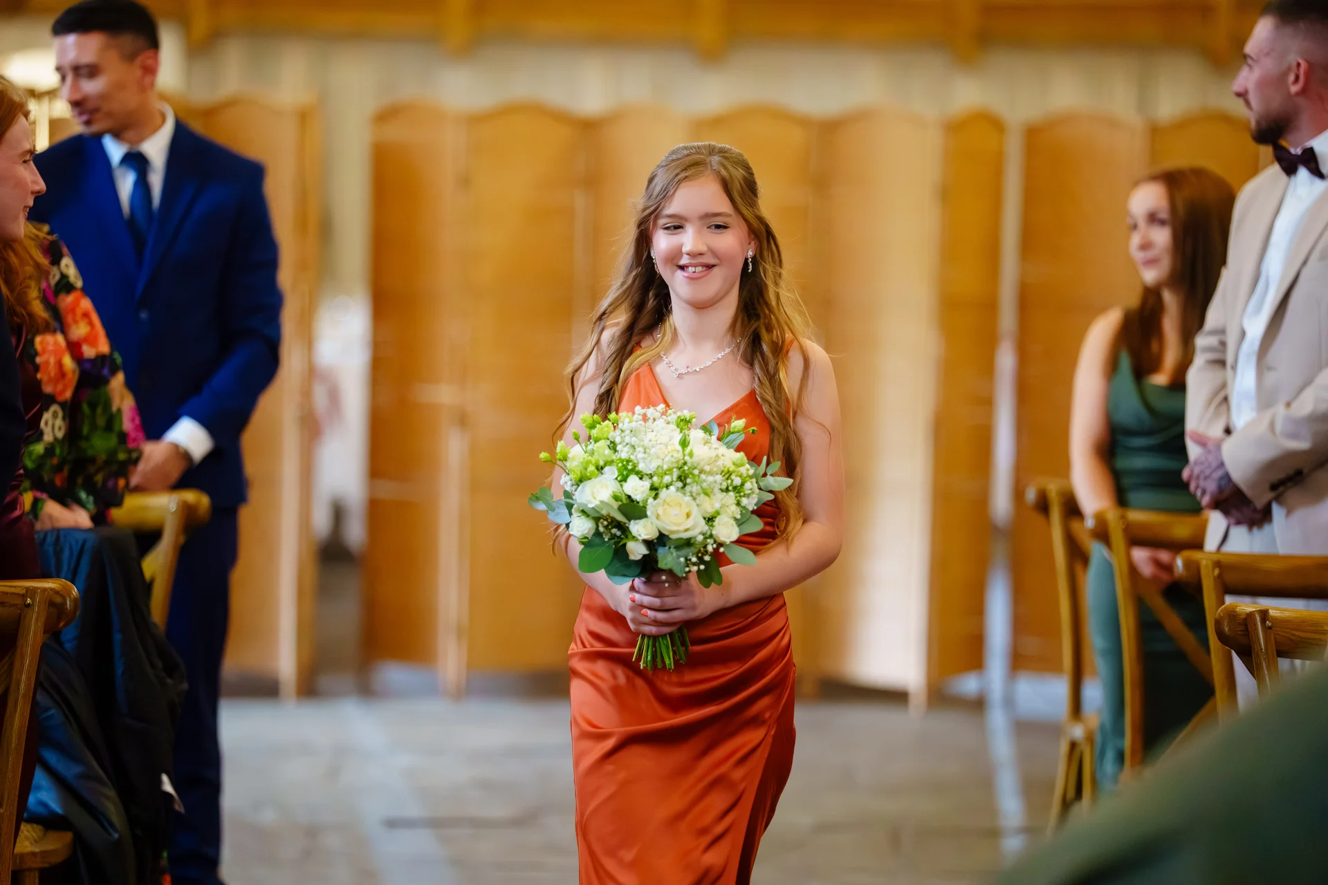 A smiling junior bridesmaid in a satin rust-orange dress walks down the aisle holding a bouquet of white roses and greenery.