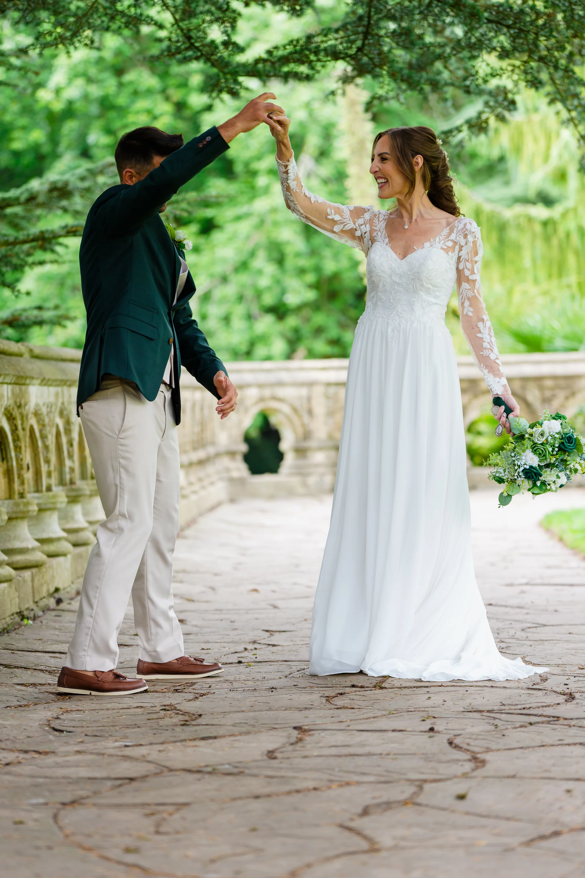 A smiling bride and groom share a joyful dance on a stone bridge outdoors, surrounded by lush green foliage and trees.