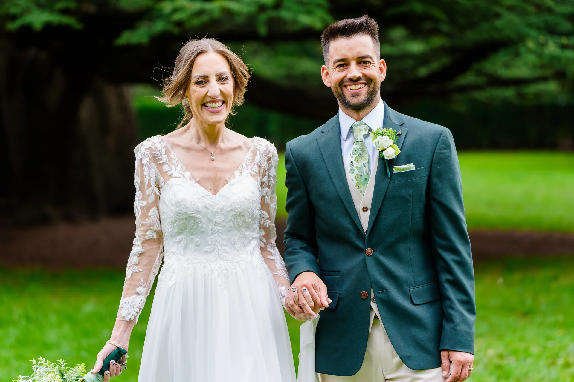 A smiling bride in a lace long-sleeve gown and groom in a dark green suit walk hand in hand outdoors on a lush green lawn.