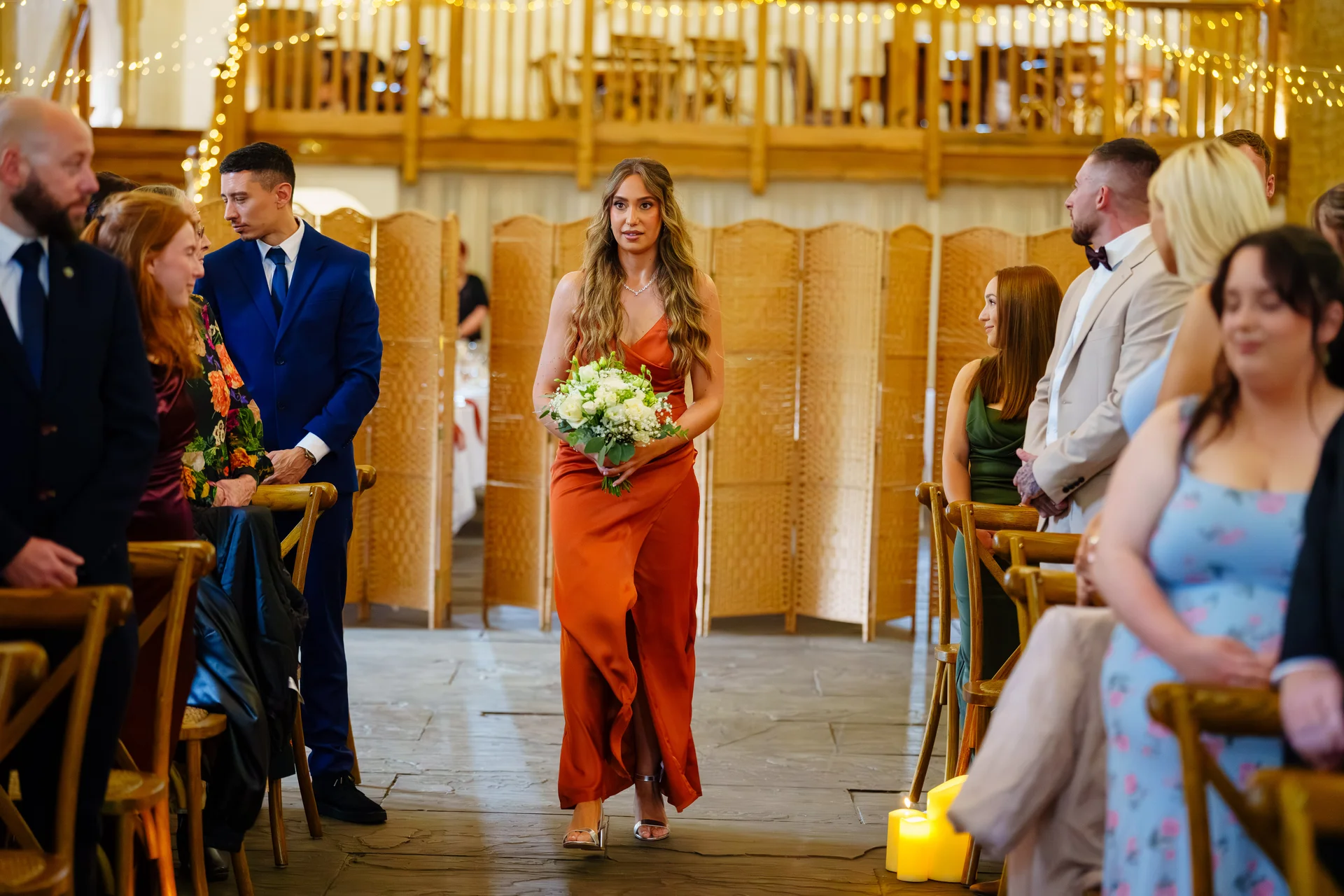 A bridesmaid in a vibrant orange dress walks down the aisle carrying a white floral bouquet during an indoor barn wedding ceremony.