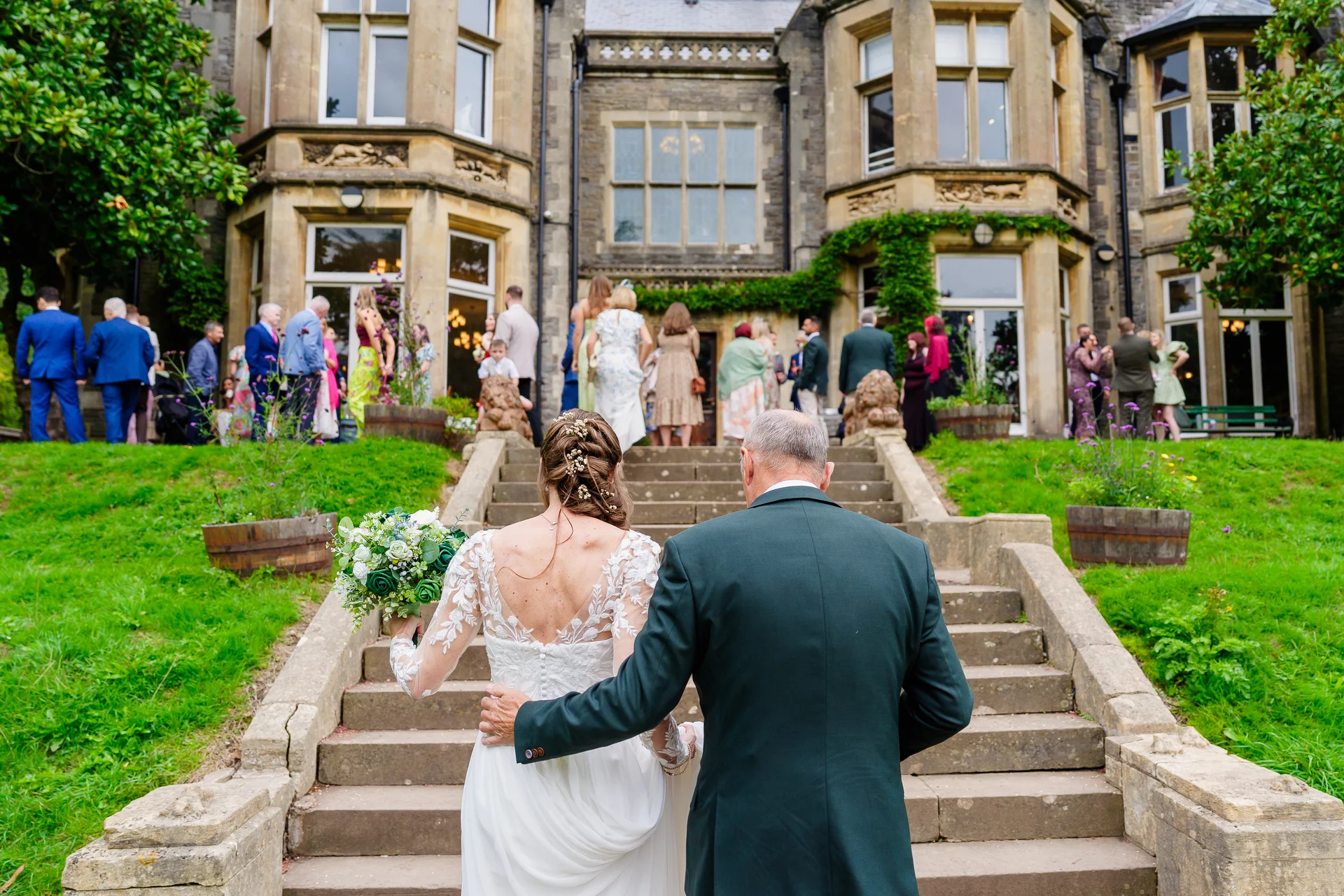 A bride in a lace gown and her father walk arm in arm up stone steps toward a grand country house venue with wedding guests in the background.