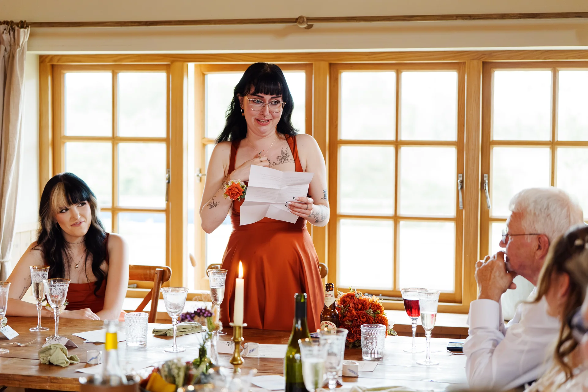 A smiling bride wearing a burnt orange dress stands at a wooden banquet table reading a speech from paper, framed by large wooden windows.