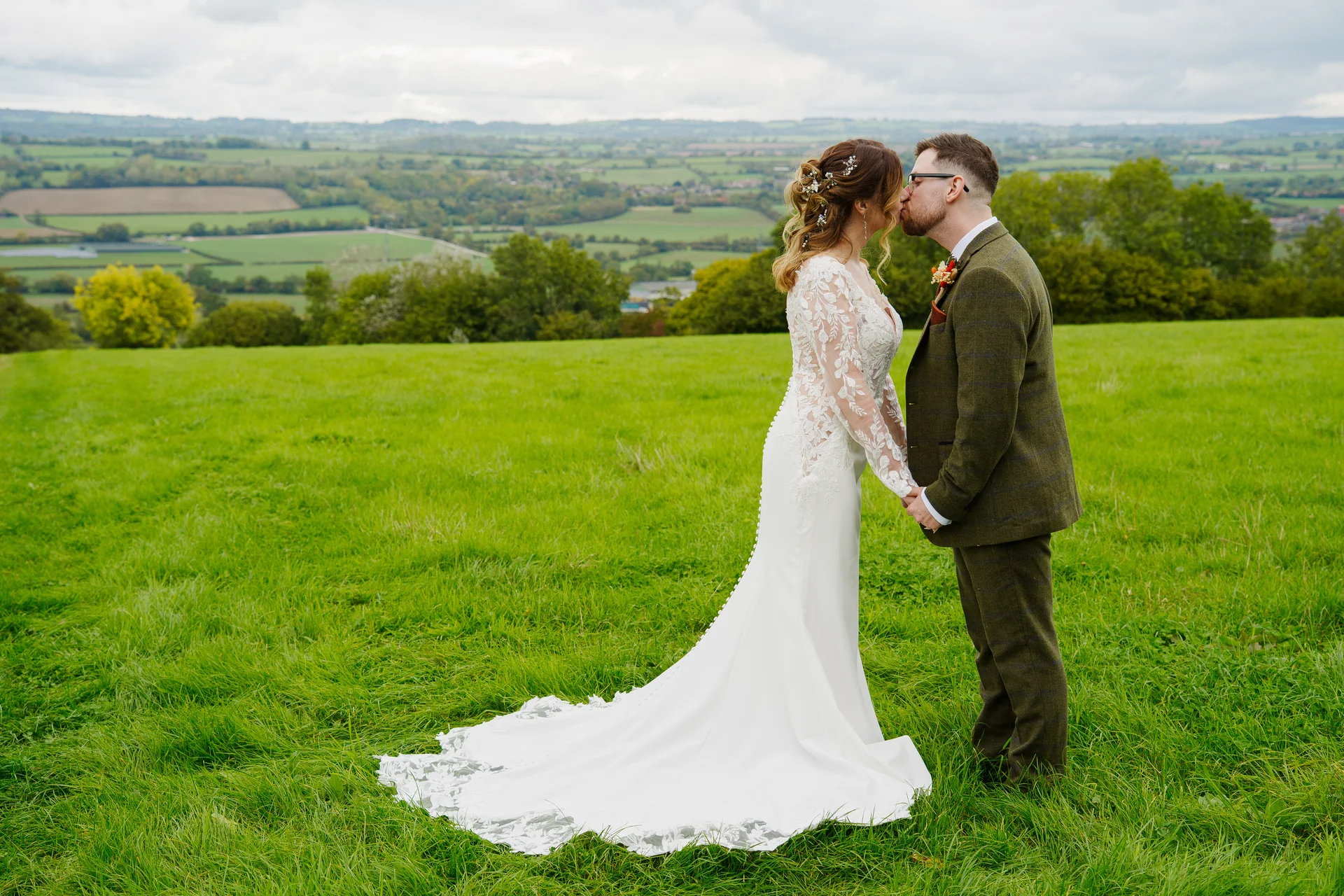 A newlywed couple shares a tender kiss while standing hand in hand on a lush green hillside overlooking a sweeping countryside view.