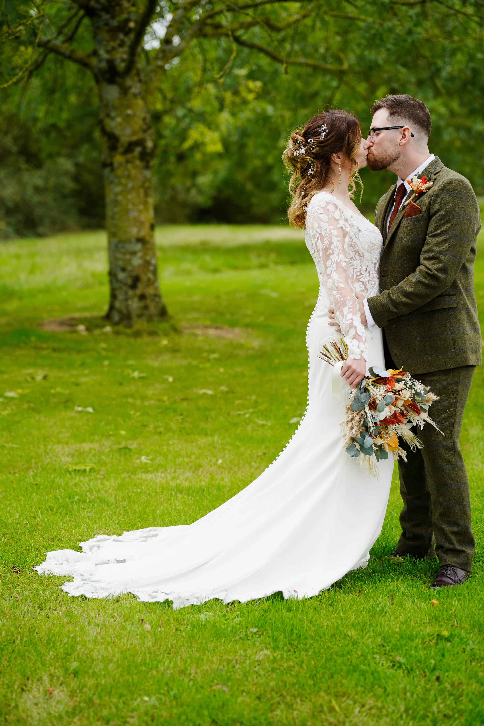 Newlyweds share a tender kiss outdoors on a vibrant green lawn, with the bride wearing a long-sleeved lace and crepe dress and holding a dried flower bouquet, while the groom wears an olive green tweed suit.