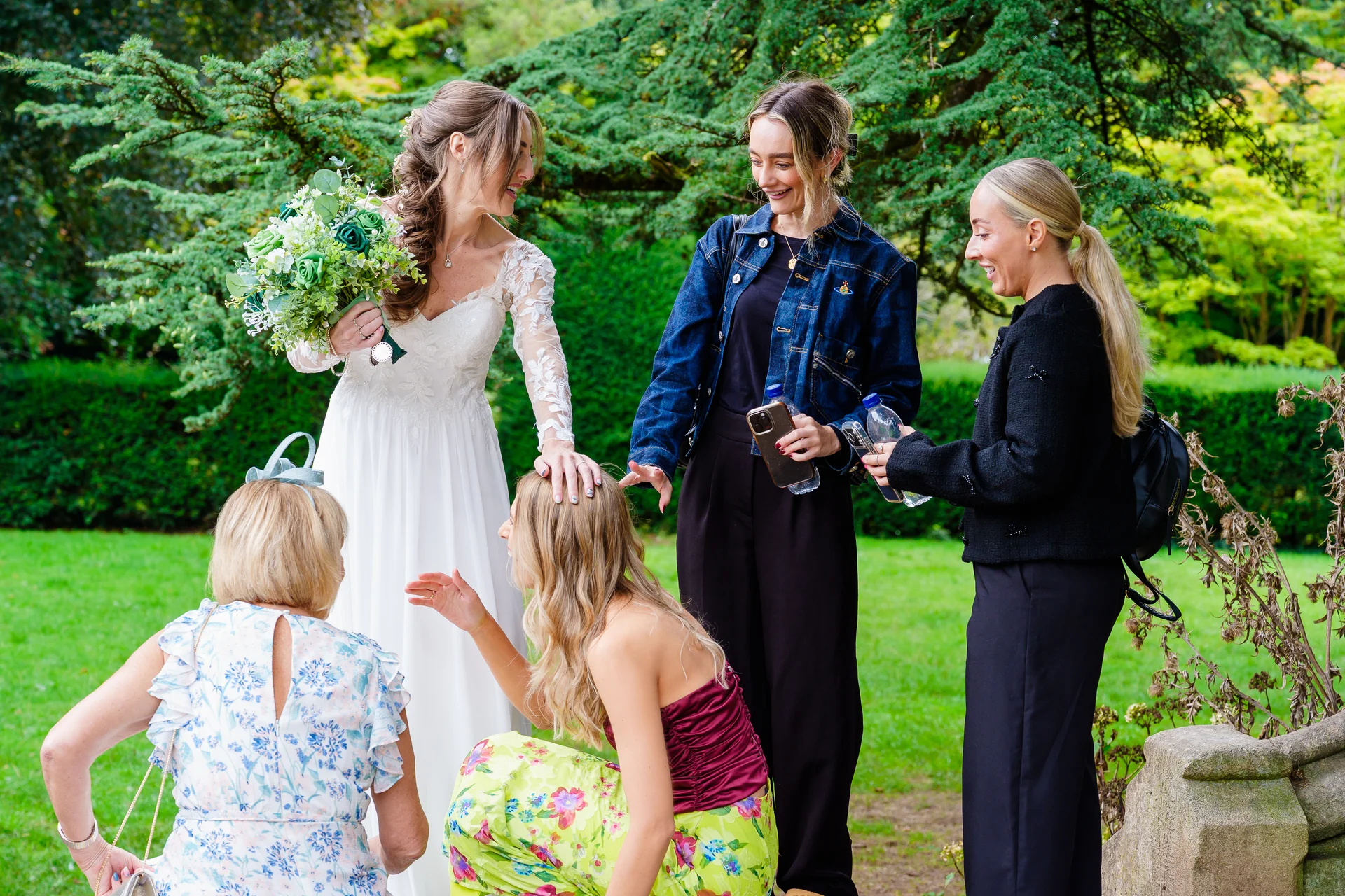 A joyful bride in a long-sleeved lace wedding dress holds a bouquet while laughing with her bridesmaids and guests in a lush green garden setting.