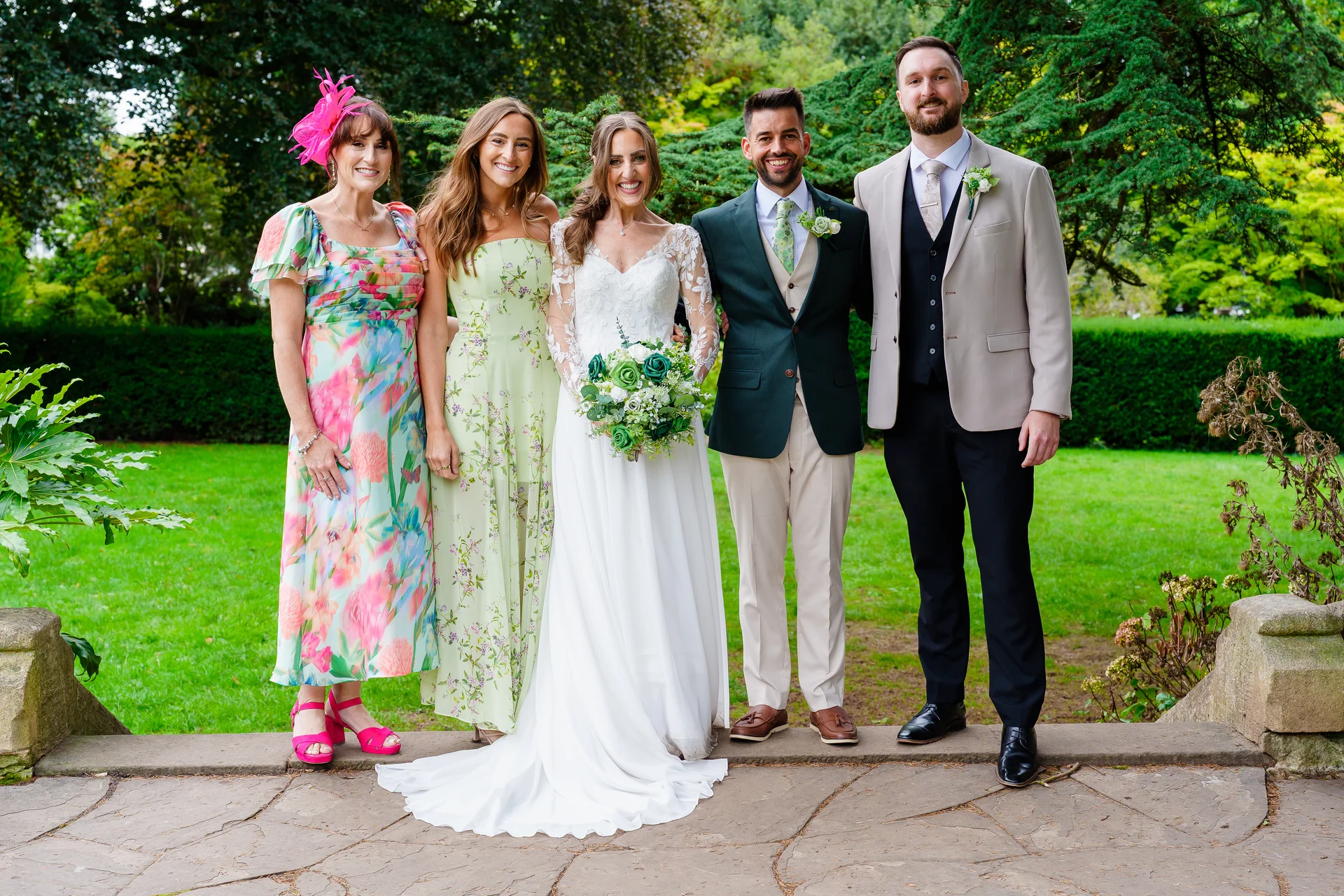 A happy wedding group stands together on a stone terrace in front of a lush green garden, featuring the bride in a white lace gown holding a bouquet alongside family members in vibrant outfits.