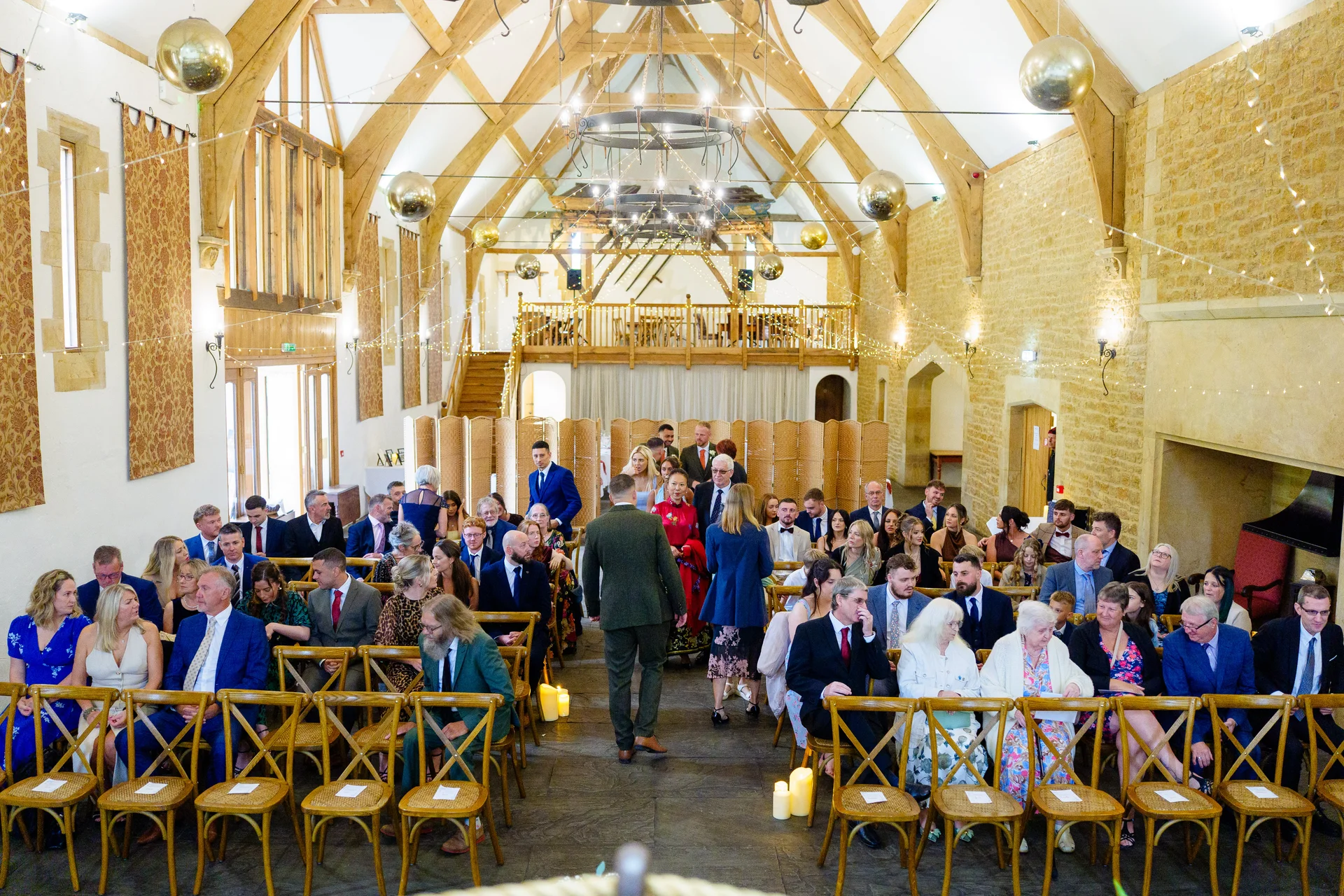 Guests are seated on wooden cross-back chairs inside a magnificent stone barn featuring high vaulted wooden beams, exposed stone walls, and a canopy of fairy lights with hanging disco balls.