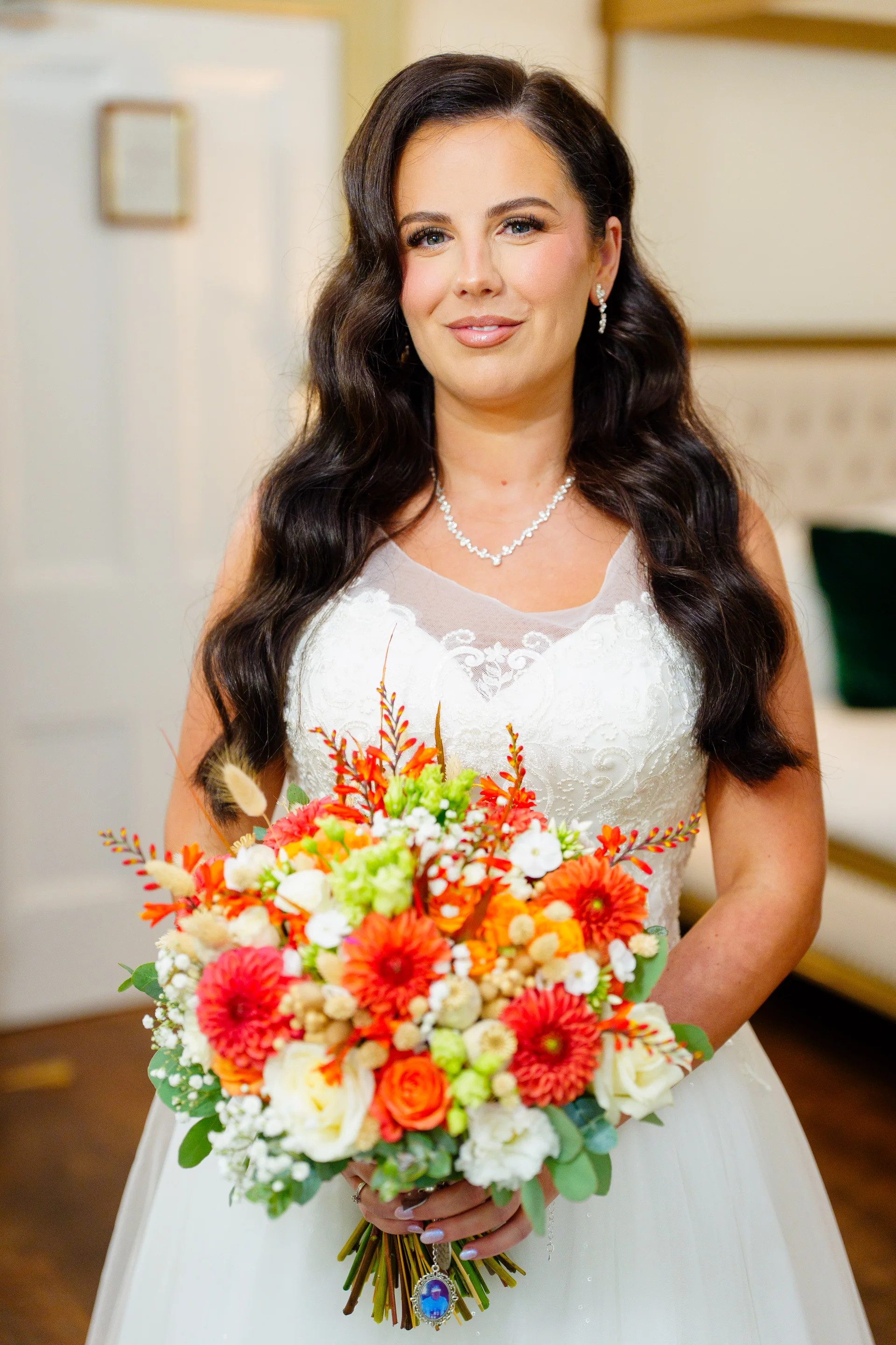 A stunning bride with dark wavy hair smiles gently while holding a vibrant, colourful hand tied bouquet featuring bright orange and red flowers.