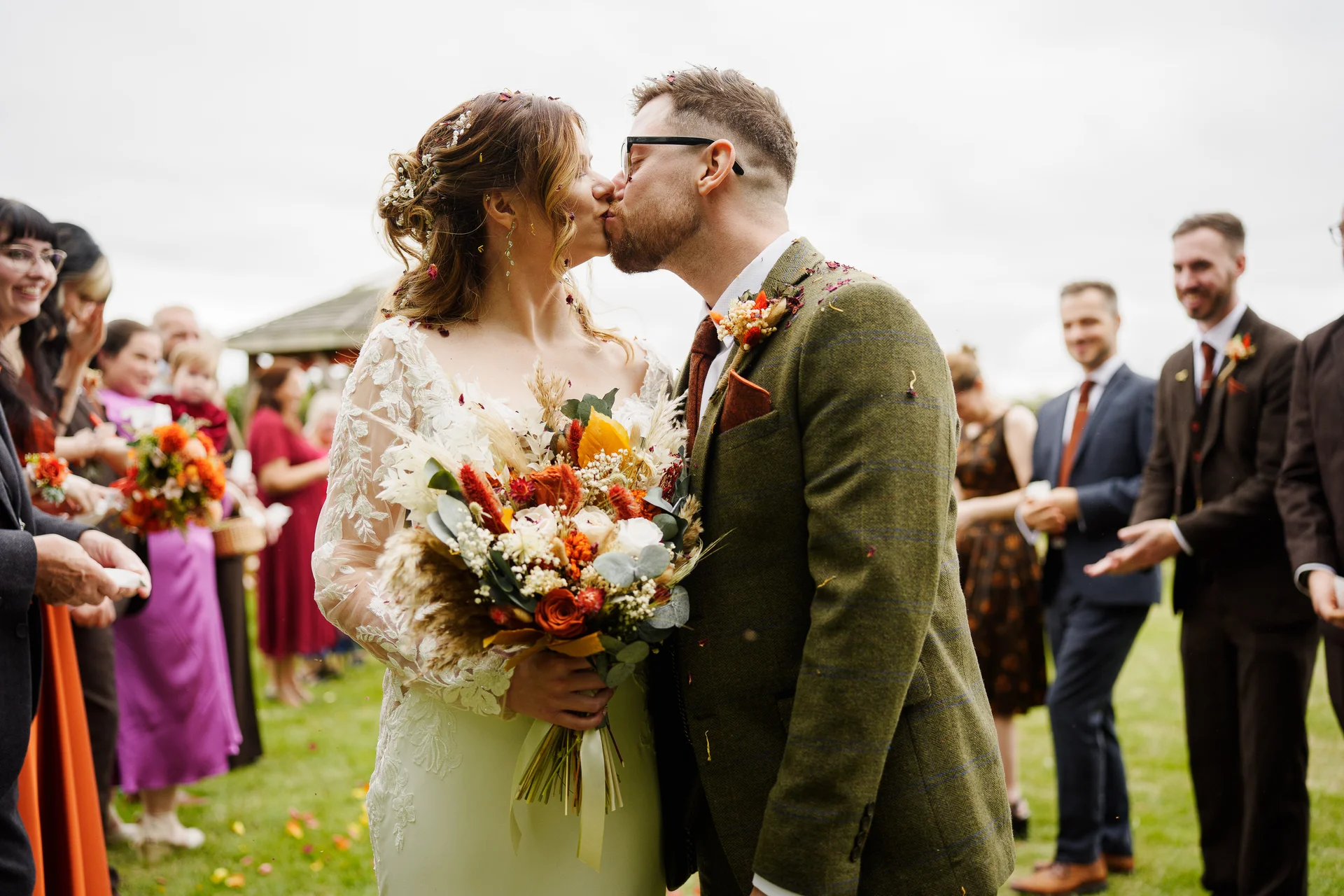 A newlywed couple share a romantic kiss surrounded by cheering guests throwing colourful confetti during an outdoor wedding ceremony.