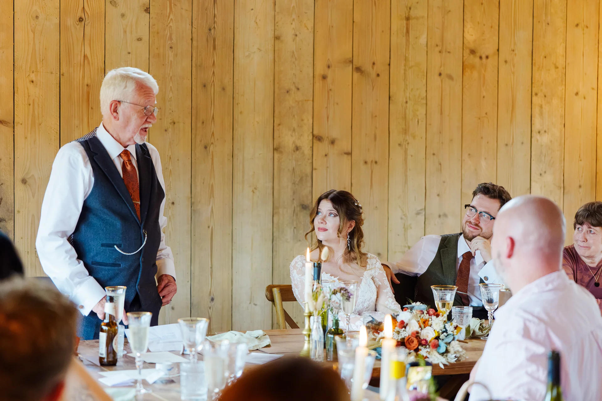 An older gentleman gives a speech at the top table inside a rustic wood-panelled barn while the bride and groom look on attentively.