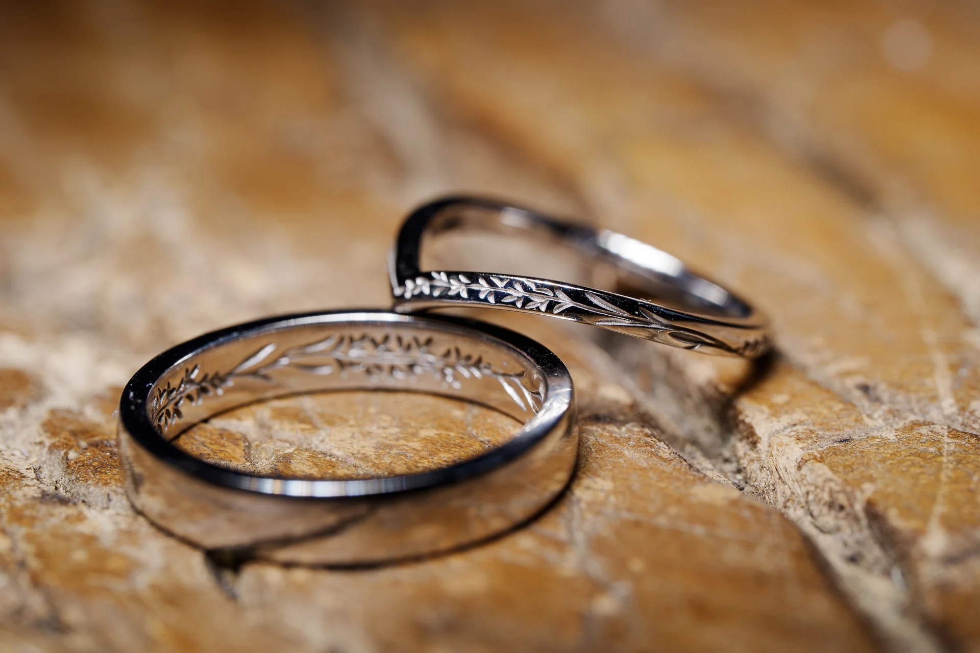A macro detail shot showcases two intricately engraved silver wedding bands resting on a textured wooden surface.