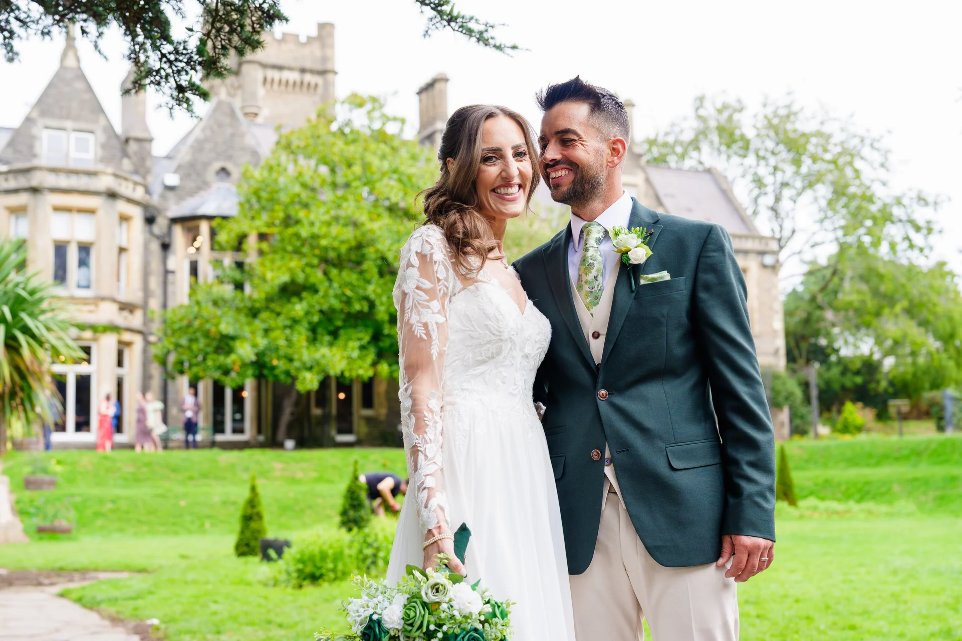 A joyful bride and groom pose smiling together in front of a grand manor house with lush green gardens on their wedding day.