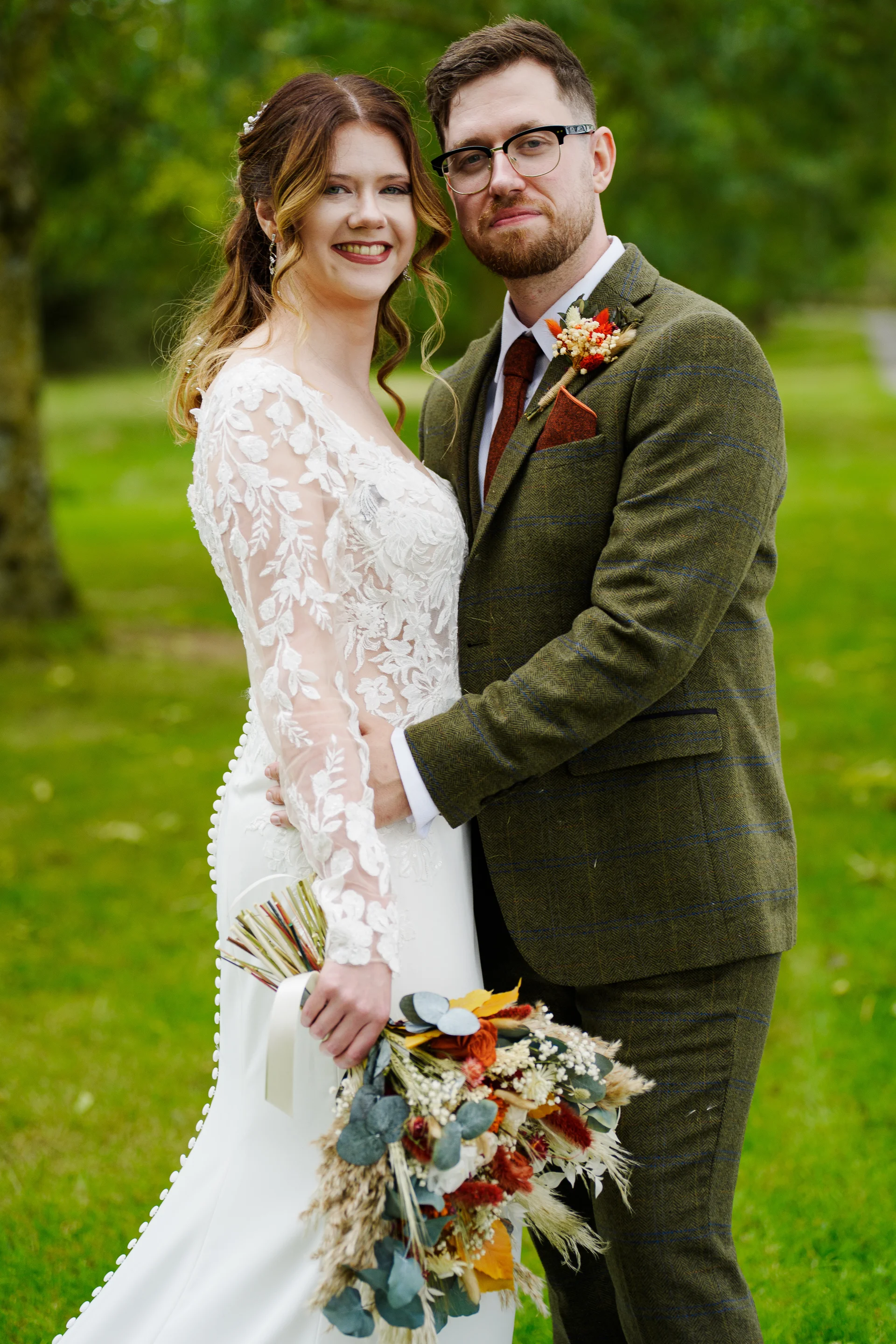 A smiling bride holding a dried flower bouquet next to the groom in a green tweed suit outdoors in a lush green setting.