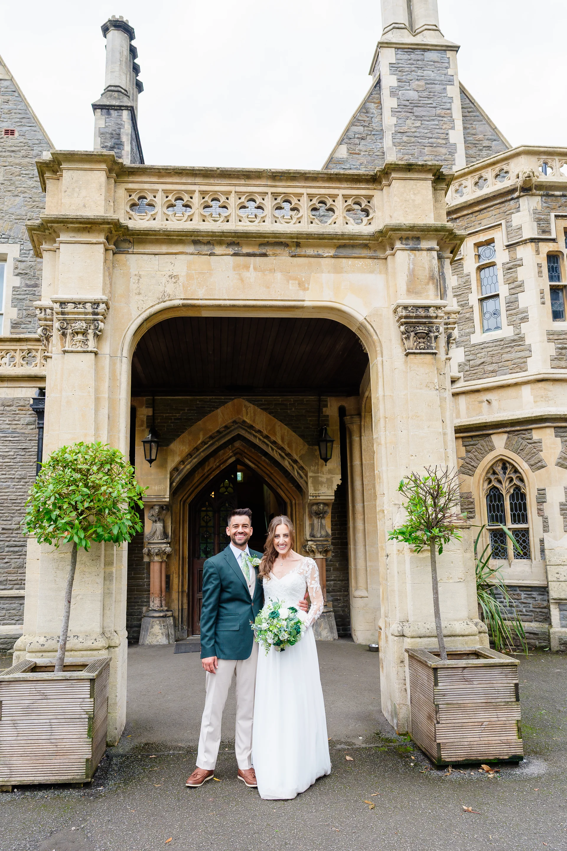 A smiling bride and groom pose together outside the grand stone entrance of a historic manor house venue.