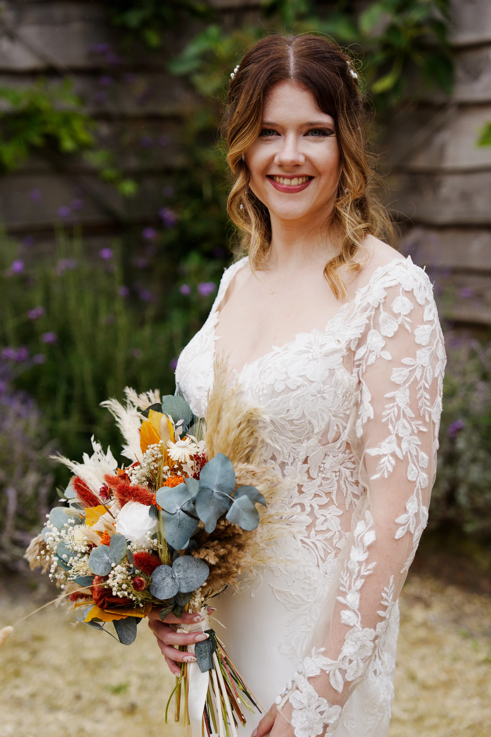 A smiling bride wearing a lace long-sleeve wedding dress and holding a vibrant autumn-toned bouquet with dried grasses and eucalyptus.