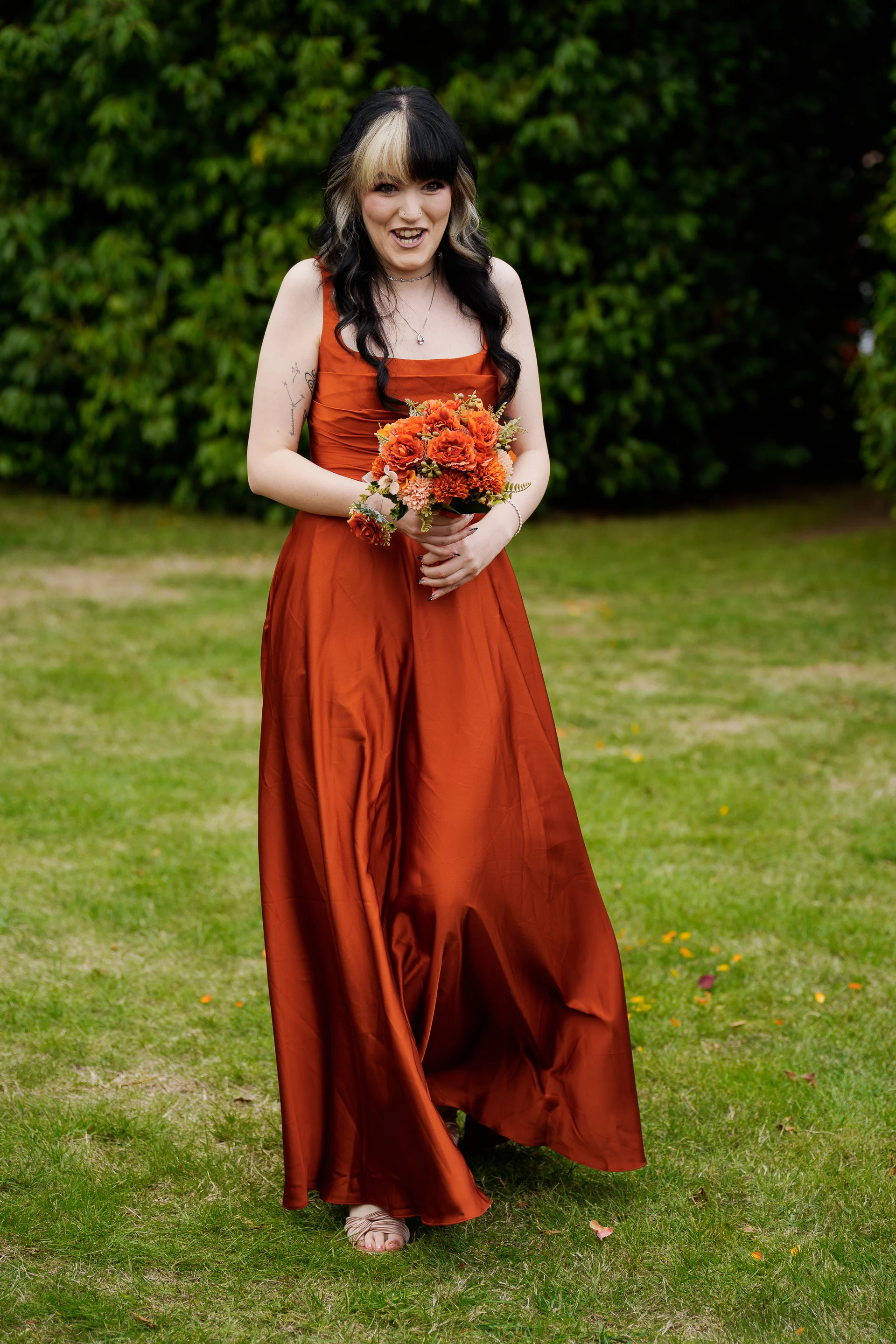 A laughing bride holding an orange bouquet stands outdoors on a green lawn wearing a flowing terracotta satin dress.