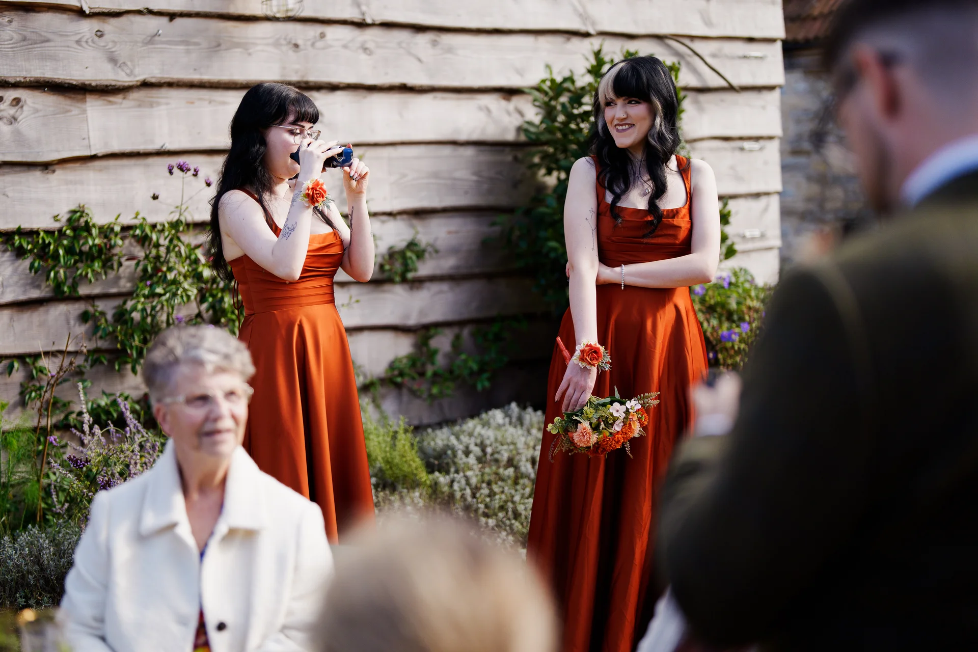 Bridesmaids wearing vibrant rust-coloured dresses smile and hold bouquets outdoors in front of a rustic wooden barn while guests look on.