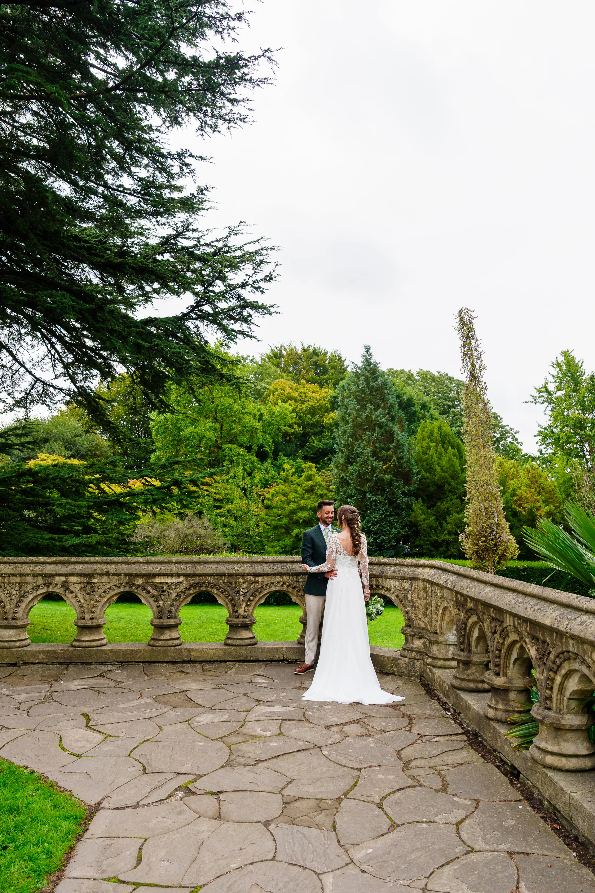 A bride in a long-sleeved lace gown and groom in a green suit share a tender glance on a paved stone terrace overlooking lush green gardens.
