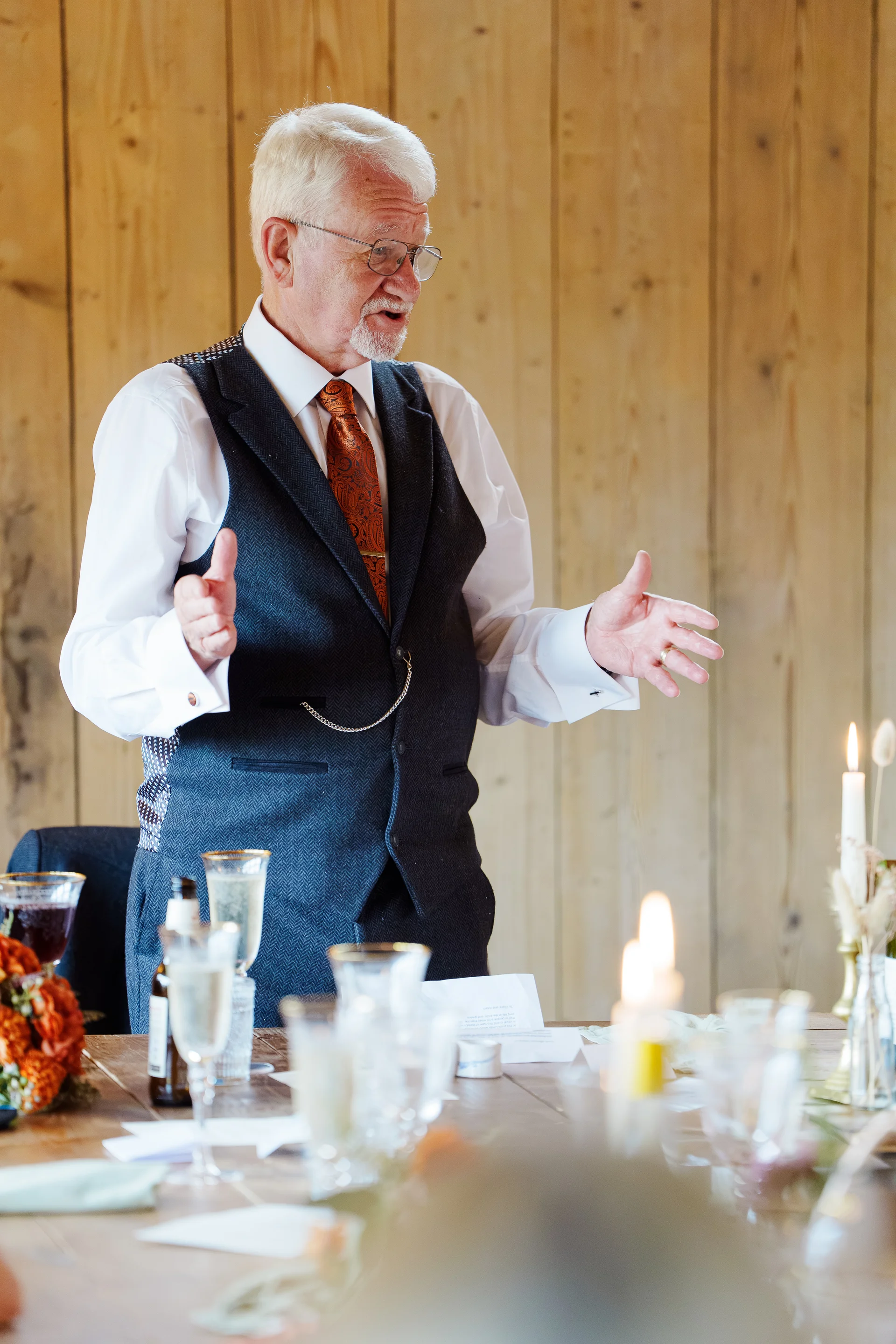 An older gentleman delivers an emotional wedding speech in a rustic barn venue, wearing a tweed waistcoat and gesturing with his hands.