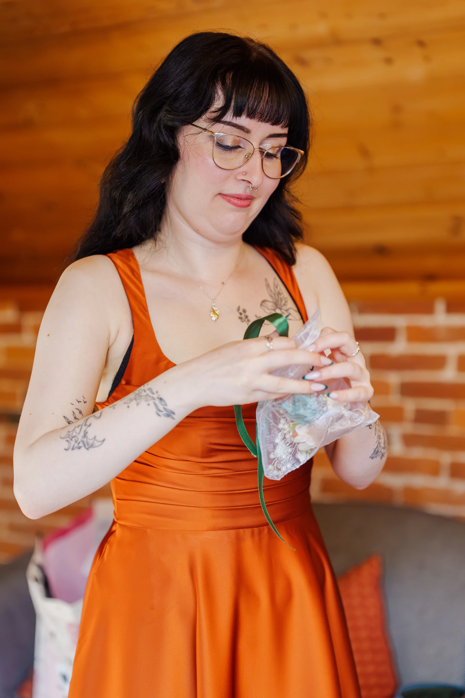 A smiling bride wearing glasses and a burnt orange dress delicately opens a small favor bag during morning preparations.