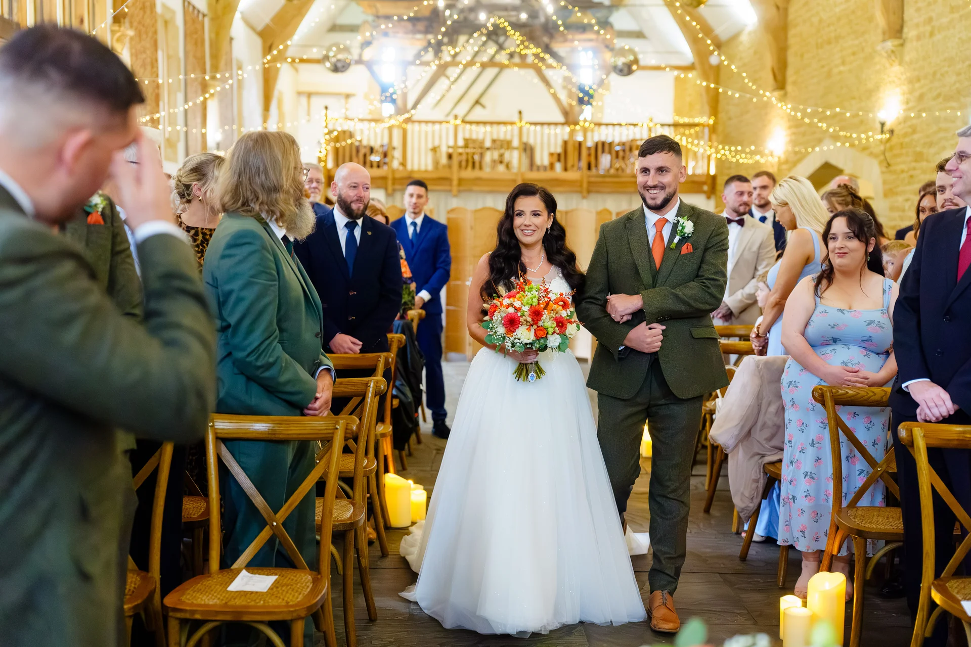 A beaming bride and groom walk hand in hand down the wooden-chaired aisle of a rustic stone barn adorned with twinkling fairy lights.